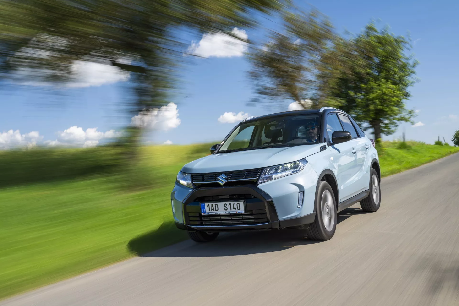 Un SUV compacto Suzuki Vitara 2024 (2ª generación con rediseño) de color azul claro en movimiento en un camino rural. La imagen captura el cuarto delantero del coche, mostrando la parrilla delantera, los faros, parte del capó, el parabrisas y el lado del conductor, incluyendo el arco de la rueda delantera, parte de la puerta trasera y una porción del arco de la rueda trasera. El coche está nítido contra un fondo y una carretera borrosos, lo que indica velocidad. Las ruedas muestran un ligero desenfoque, lo que sugiere rotación. Iluminado por luz natural brillante en un día soleado, arrojando sombras en la parte inferior del vehículo, la imagen resalta elementos de diseño como los pliegues del capó, el diseño del parachoques delantero y el patrón de la parrilla. Los faros LED son visibles, lo que podría indicar características modernas como sensores de estacionamiento. La matrícula es visible sin información sensible.