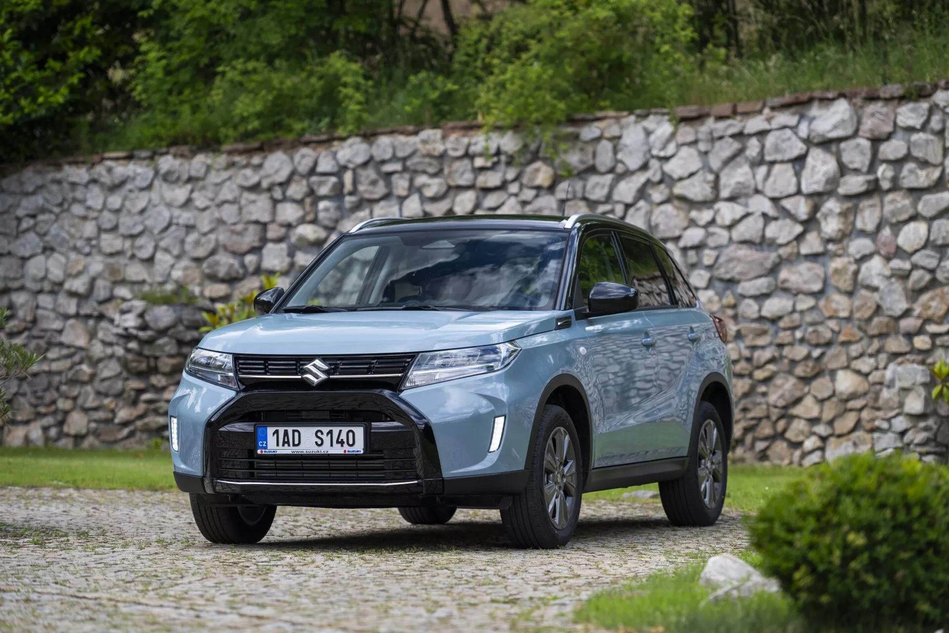 Fotografía de alta calidad de un coche Suzuki en un entorno al aire libre, capturada desde un ángulo frontal de tres cuartos cerca de zonas verdes o un parque. La vista incluye la parrilla delantera con una insignia prominente de Suzuki, faros de aspecto moderno posiblemente con tecnología LED, luces antiniebla dentro del parachoques y el perfil del lado del conductor con ruedas visibles, puerta y espejos laterales. El coche está sobre una superficie de grava con una pared de piedra en el fondo, enfatizando el ambiente rústico y natural. La escena está suavemente iluminada por la luz dispersa, con poca profundidad de campo, desenfocando el fondo para enfocar en el diseño y detalles tecnológicos del coche.