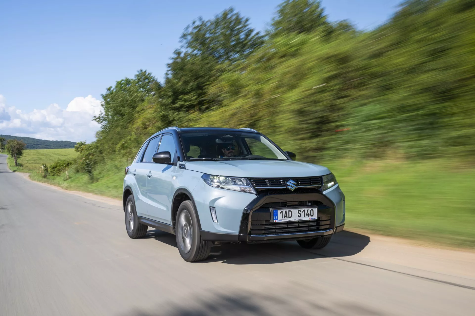 Un SUV moderno, el Suzuki Vitara 2024 (segunda generación, facelift 2) mostrado en movimiento desde un ángulo bajo y tres cuartos frontal. La imagen destaca el distintivo frente del vehículo, que incluye una parrilla prominente con listones horizontales, faros LED afilados y angulares, y una llanta de aleación de múltiples radios. El SUV se captura conduciendo en una carretera de asfalto con un paisaje verde natural en el fondo, con un desenfoque de movimiento que transmite velocidad y dinamismo. Las características adicionales incluyen un capó inclinado, espejos aerodinámicos para mejorar la aerodinámica, mayor altura desde el suelo y un interior espacioso, todo acentuado por un acabado de pintura metálica.