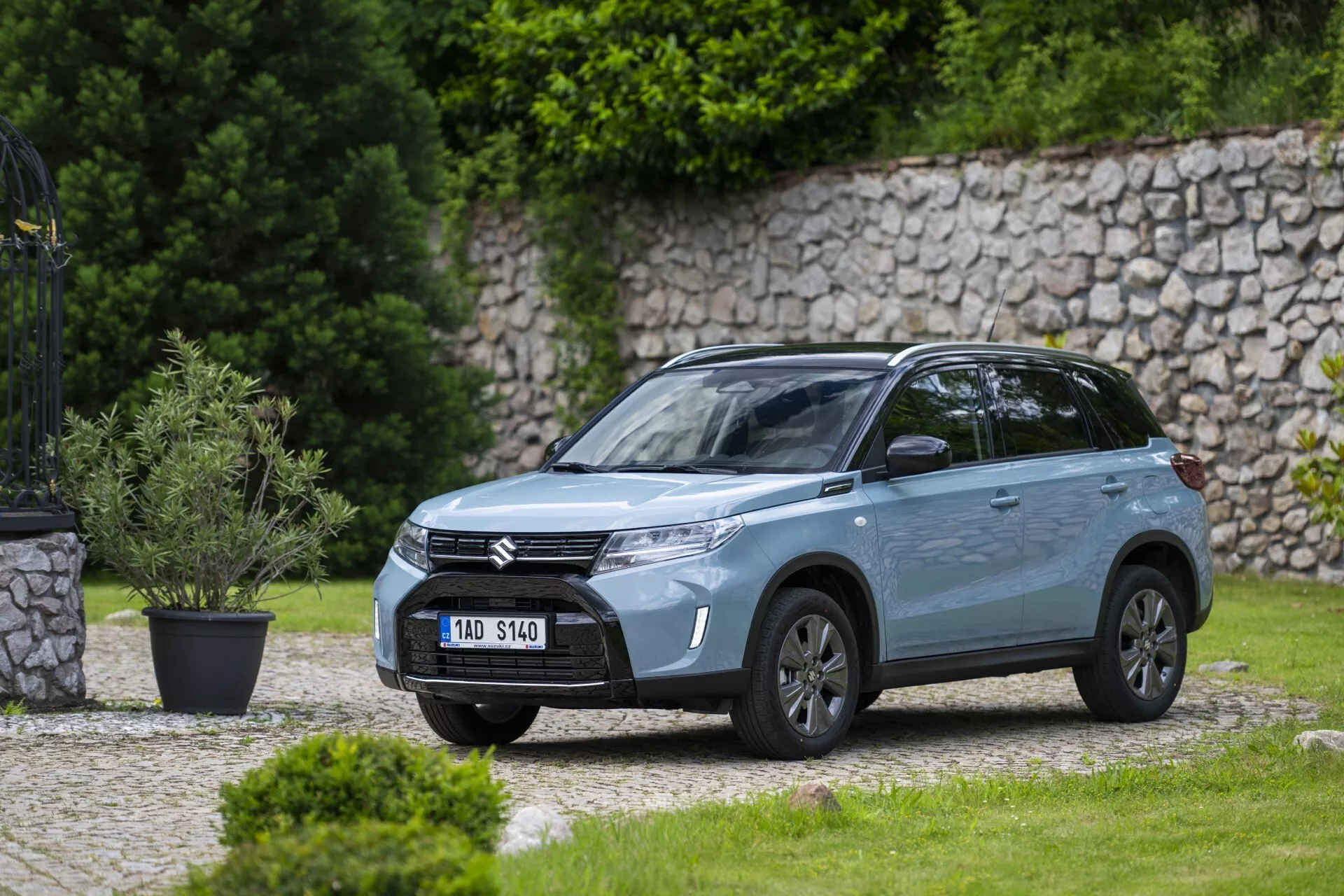 Vista frontal lateral en ángulo de un moderno SUV Suzuki Vitara 2024 estacionado al aire libre en un área residencial o similar a un parque con un césped bien cuidado y un muro de piedra en el fondo. La imagen, tomada desde una distancia media, muestra la parrilla frontal con barras horizontales con el logotipo de la compañía, faros LED elegantes con luces diurnas, parachoques esculpido con luces antiniebla integradas, llantas de aleación, espejos laterales con señales integradas, acabado de pintura metálica, líneas de carrocería dinámicas, ventanas tintadas, rieles en el techo y antena en el techo, destacando su diseño y características modernas.