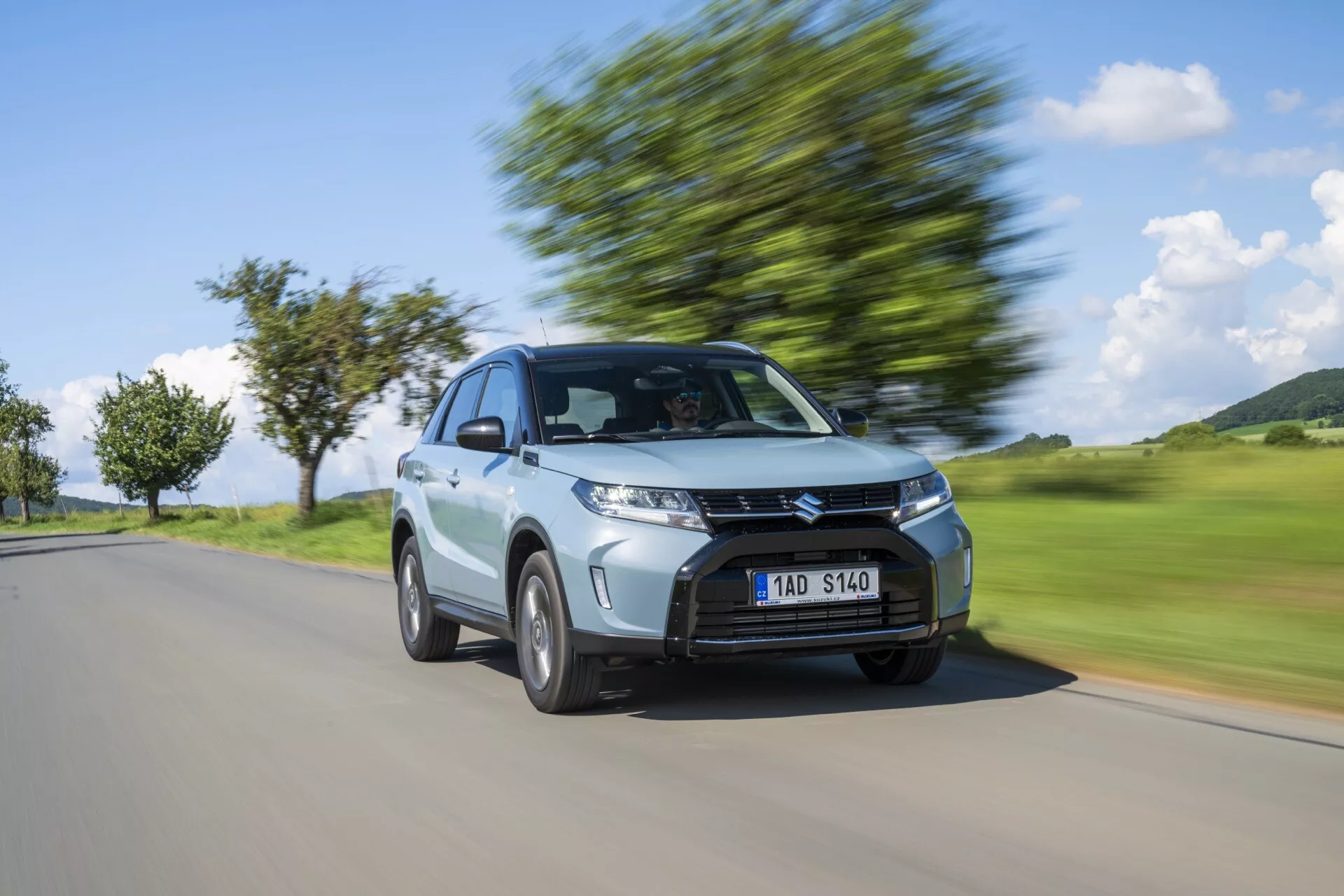 Imagen dinámica de un automóvil hatchback plateado Suzuki Vitara 2024 en movimiento capturado desde un ángulo frontal de tres cuartos, destacando su diseño moderno con una parrilla prominente, faros angulares, techo inclinado y ruedas de aleación en movimiento con desenfoque de fondo que indica velocidad.