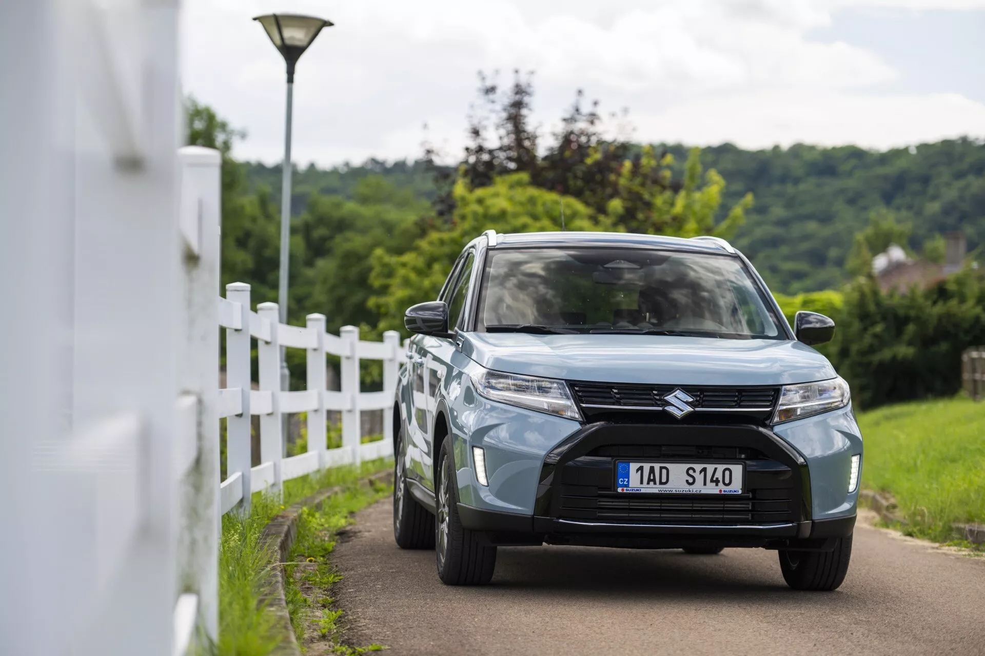 Escena diurna al aire libre que muestra el Suzuki Vitara 2024 (facelift de 2da generación) capturado desde una perspectiva tres cuartos frontal. La imagen enfatiza los elementos de diseño del vehículo incluyendo la parrilla delantera prominente con diseño de listón horizontal y la insignia central, faros modernos LED o HID, luces antiniebla empotradas en el paragolpes inferior, y el color azul claro o gris. El coche está conduciendo por un carril estrecho cerca de una cerca blanca con un fondo ligeramente borroso, lo que sugiere una pequeña profundidad de campo. También se observan ruedas de aleación, espejos laterales con señales de giro integradas y paragolpes delantero parcialmente visible con entrada de aire central.