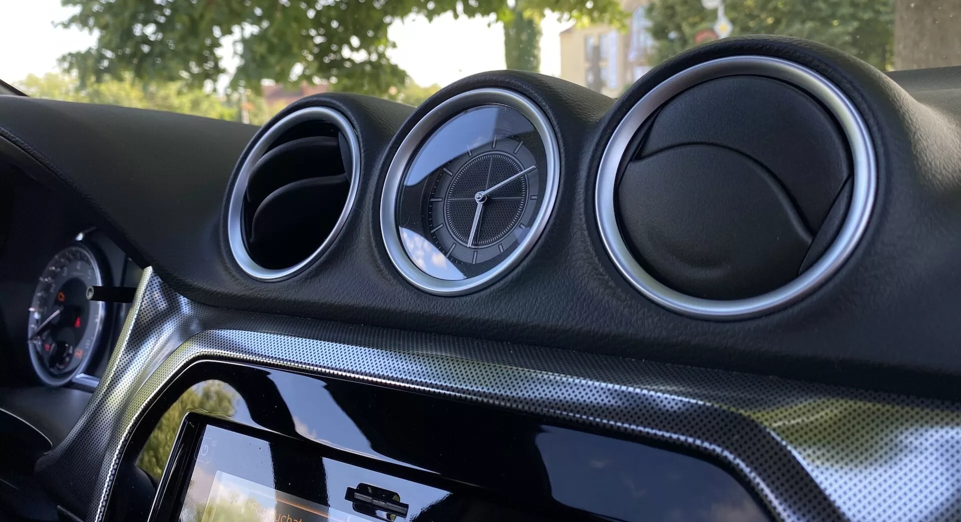 Vista interior de cerca del Suzuki Vitara 2018 enfocada en el tablero central. El tablero cuenta con tres ventilaciones circulares de aspecto metálico con el emblema del fabricante, un sistema de infoentretenimiento con pantalla táctil y borde negro brillante, material con apariencia de fibra de carbono y iluminación ambiental para mejorar la estética.