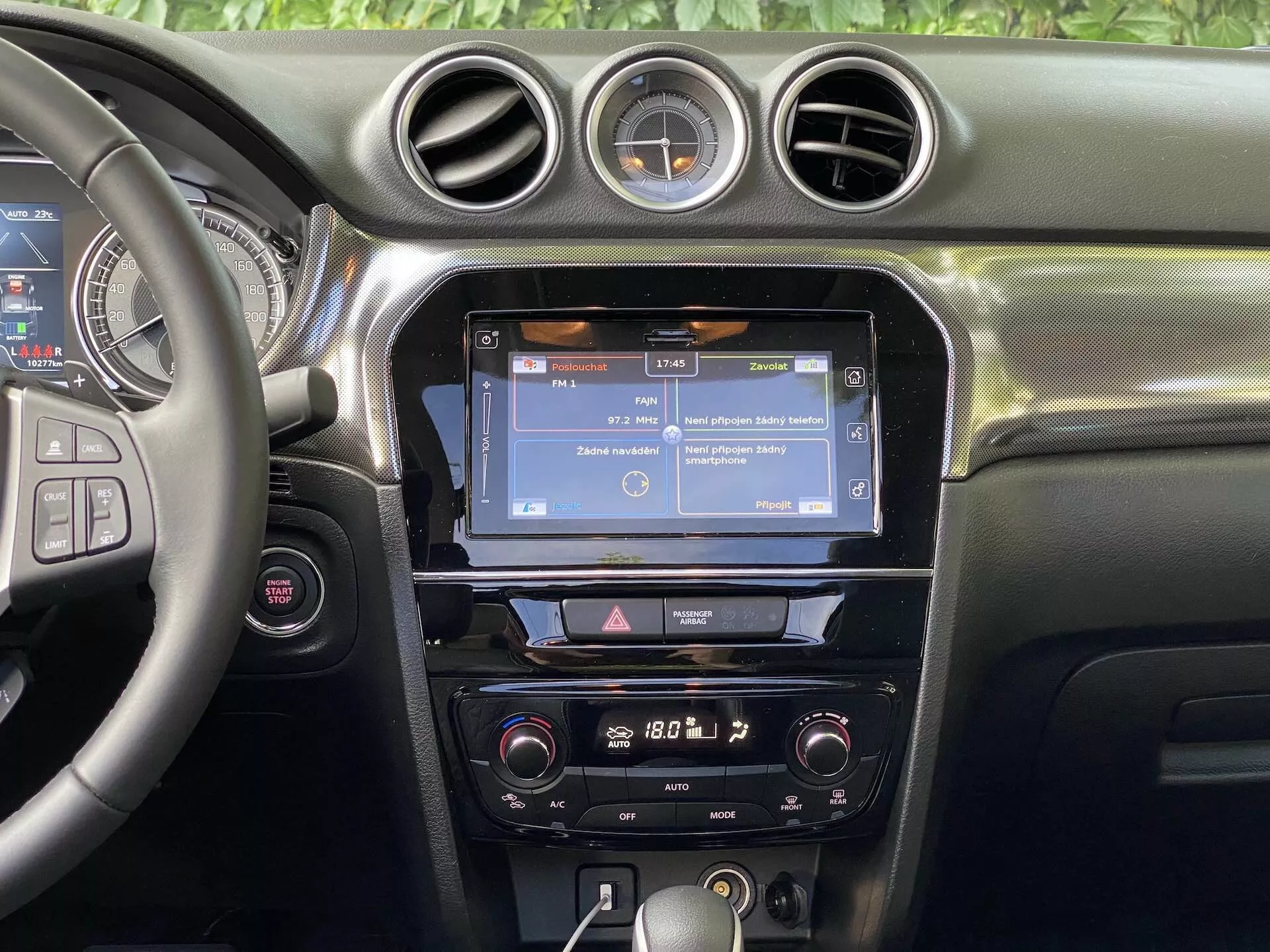 Vista interior del Suzuki Vitara 2018 enfocándose en el tablero central. La sección muestra un volante parcialmente visible a la izquierda con múltiples botones de control, probablemente para el audio, llamadas manos libres y control de crucero. La consola central muestra una pantalla táctil de infoentretenimiento, posiblemente mostrando navegación o una interfaz de menú, flanqueada por ventilaciones verticales y rematada con tres salidas de aire circulares. Debajo de la pantalla táctil, hay una fila de botones y diales que proporcionan acceso rápido a los ajustes del infoentretenimiento y del vehículo. Los controles de clima están más abajo, sugiriendo un sistema de control de clima de doble zona. El tablero muestra un material de tacto suave con costuras visibles, y un reloj analógico redondo está montado entre las ventilaciones circulares centrales. La visibilidad se centra en las características del tablero, sin ventanas ni parabrisas a la vista.