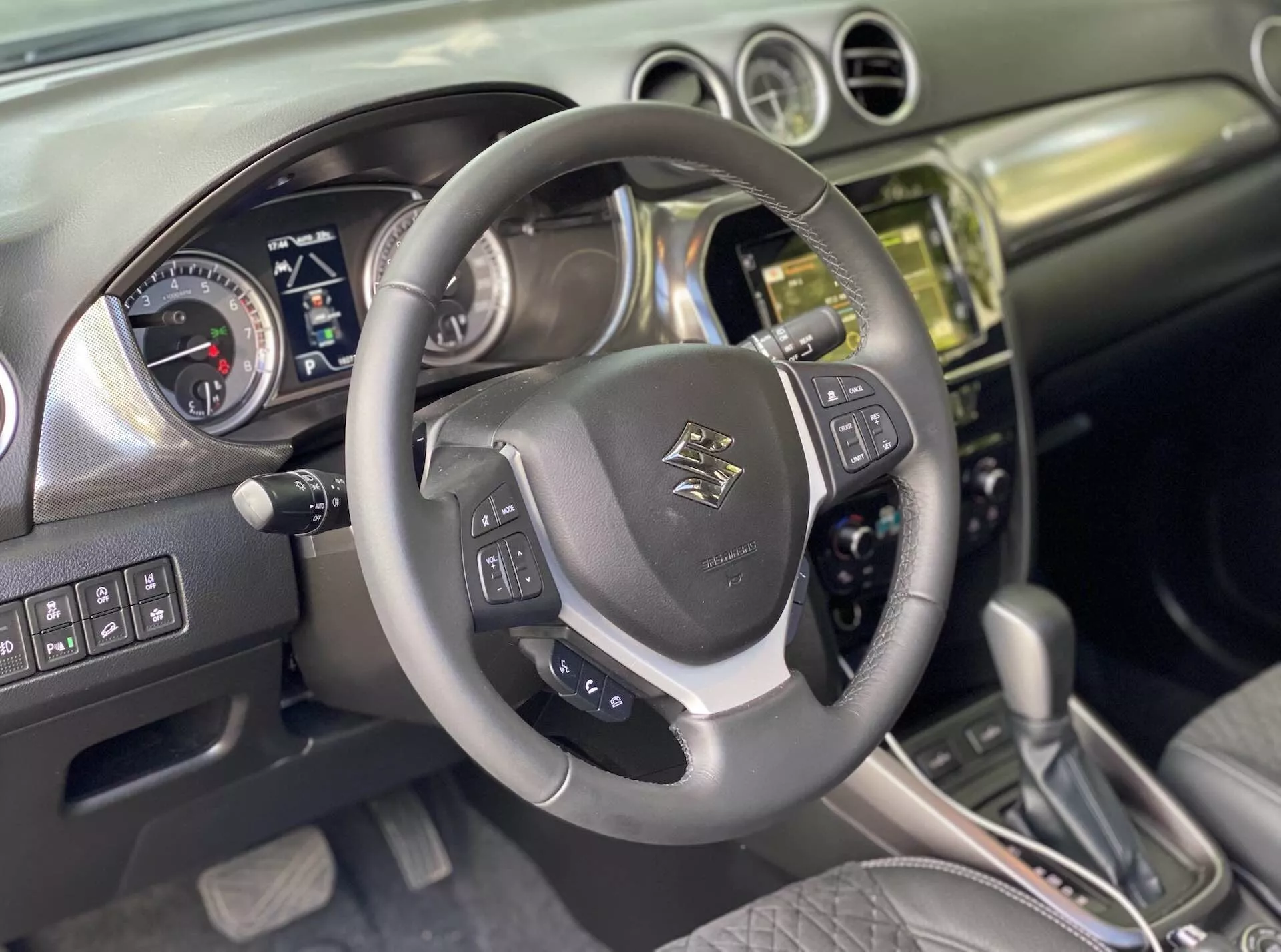 Interior del Suzuki Vitara 2018, capturando la perspectiva del conductor. Primer plano de un volante multifunción envuelto en cuero con el logotipo en el centro, controles montados para audio, control por voz y control de crucero con acentos metálicos. Detrás, un panel de instrumentos con diales analógicos para velocímetro y tacómetro, y una pantalla digital para información del vehículo, sugiere que el coche está apagado o en modo accesorio. La consola central muestra una pila terminada en metal o cromo con un sistema de infoentretenimiento de pantalla táctil, botones y perillas para controles de clima y del vehículo. La palanca de cambios automática con acentos de cuero sugiere varios modos de conducción. El interior combina cuero y metal cepillado, reflejando un diseño moderno. Se infieren características avanzadas como Bluetooth, navegación, asistentes de aparcamiento y sistemas de asistencia al conductor.