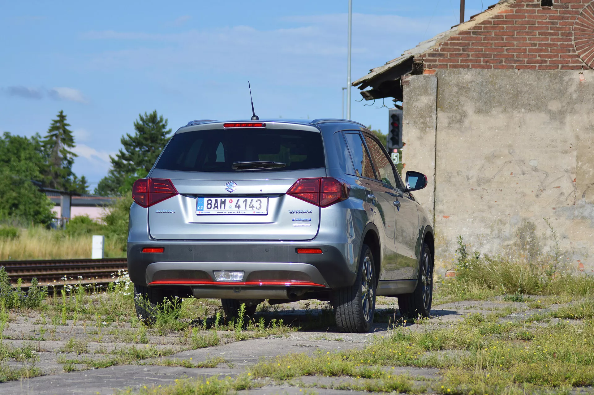 Un SUV compacto Škoda Yeti de color gris metálico estacionado en un terreno descuidado con parches de hierba y concreto, fotografiado desde una vista trasera de tres cuartos. La imagen muestra el diseño trasero distintivo del vehículo con luces traseras convencionales, limpiaparabrisas trasero, luz de freno montada en alto y parte de un alerón trasero. El logo de Škoda está centrado en el portón trasero, el nombre del modelo 'Yeti' a la izquierda y una insignia '4x4' a la derecha, indicando tracción en las cuatro ruedas. El coche tiene llantas de aleación de cinco radios, parece estar en buen estado y se encuentra en una zona industrial con vías de tren fuera de uso y una estructura en ruinas en el fondo, bajo luz solar natural.