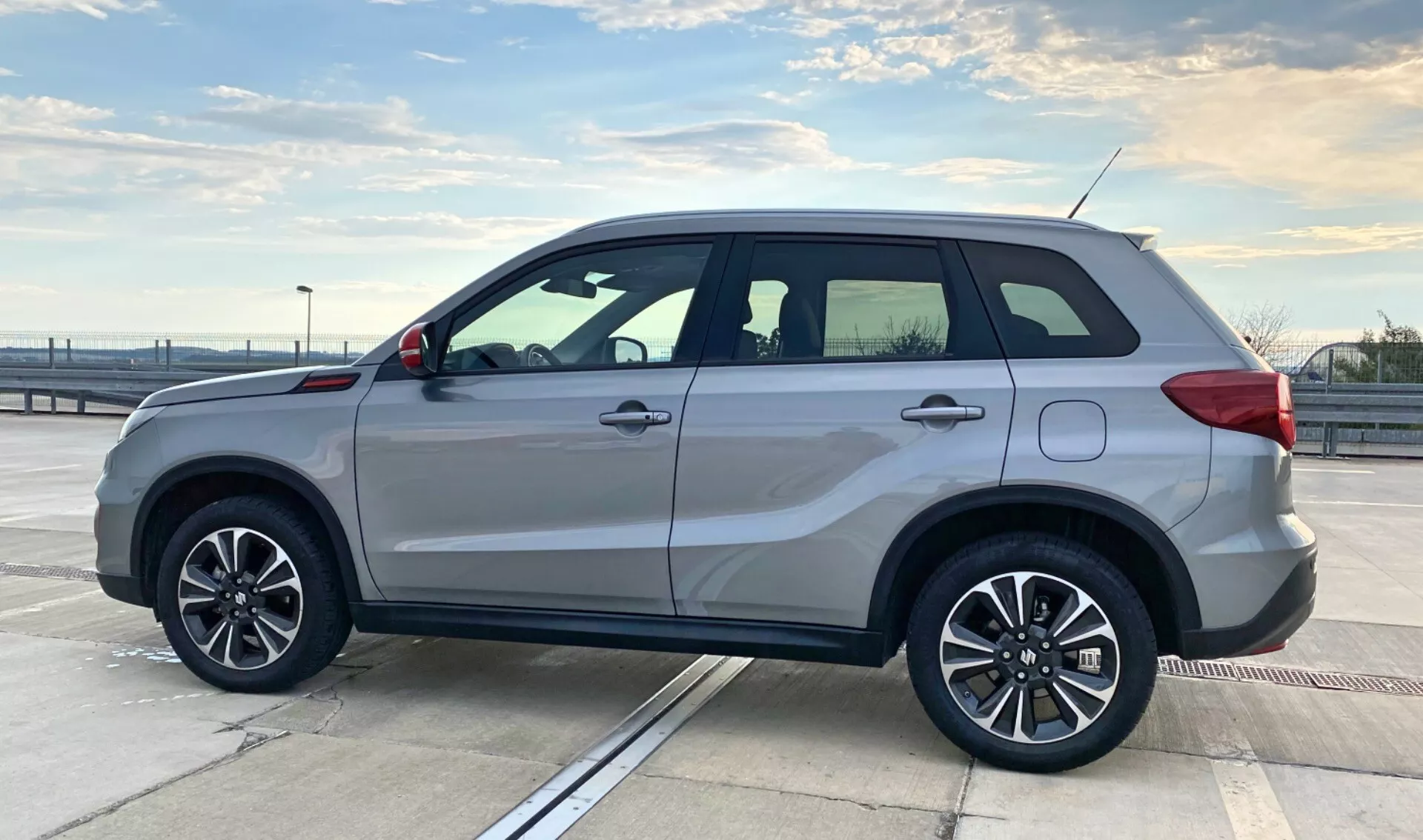 Vista lateral del Suzuki Vitara 2018 estacionado en la cubierta superior de un aparcamiento bajo un cielo despejado. El SUV presenta llantas de aleación de cinco radios, pintura plateada, señales de giro integradas en los retrovisores laterales, rieles en el techo, manijas de las puertas del mismo color que la carrocería, un sutil escalón lateral y ventanas tintadas con moldura cromada. El diseño del vehículo resalta la elegancia deportiva y la utilidad, con un cuerpo cuadrado que maximiza el espacio interior. El entorno al aire libre y la composición destacan el estilo y la construcción del automóvil, adecuado para categorías relacionadas con vehículos.