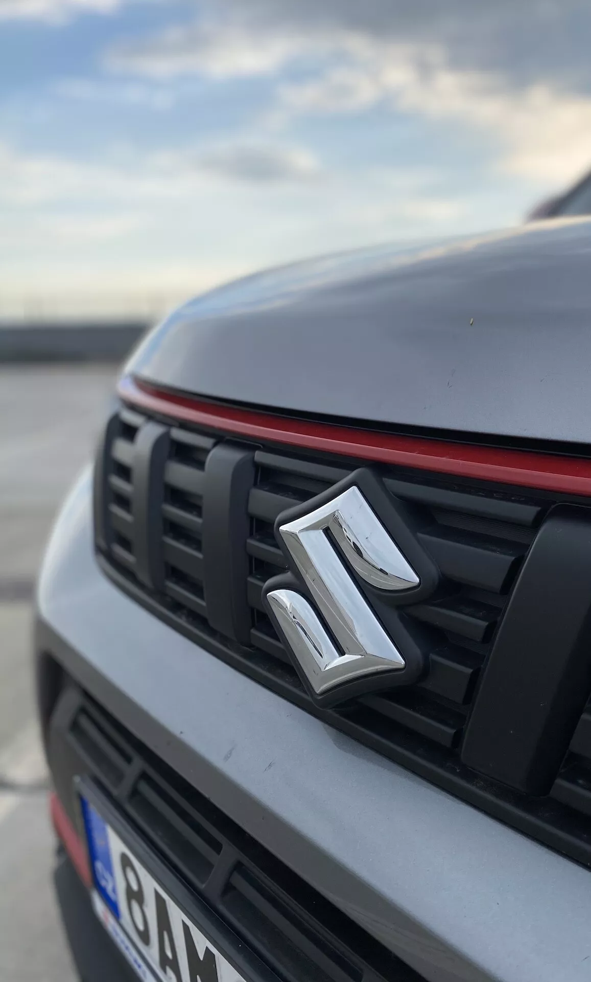 Vista de cerca y en ángulo bajo de la sección frontal de un Suzuki Vitara 2018. La imagen destaca el emblema del fabricante centrado en la parrilla, que tiene láminas horizontales y un borde rojo a lo largo del borde superior, lo que sugiere un paquete de diseño deportivo. El capó y las secciones del automóvil están desenfocados, resaltando un acabado blanco o de color claro con un pliegue discernible. El fondo también está desenfocado, insinuando un entorno al aire libre, posiblemente en un estacionamiento, durante la mañana temprano o la tarde.