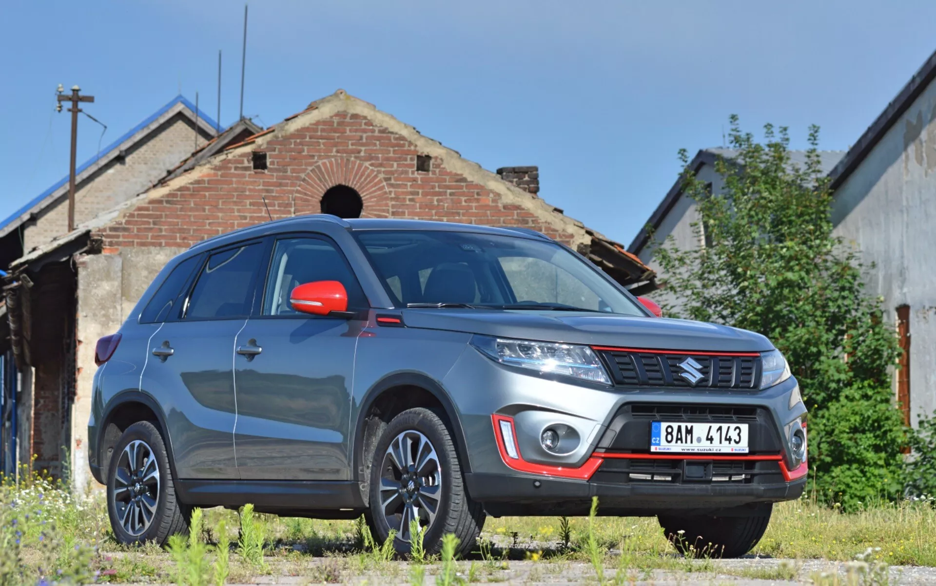 La imagen muestra un SUV Suzuki Vitara 2018 desde una vista frontal de tres cuartos bajos, destacando el lado delantero derecho del vehículo con notable distancia al suelo. Las características visibles incluyen un parachoques delantero robusto con luces antiniebla y placa de deslizamiento plateada, el emblema de la marca en la rejilla con listones horizontales, líneas definidas del capó, faros modernos posiblemente con tecnología LED o HID, ruedas de aleación multirradio, espejos laterales de color rojo con señales de giro integradas, guardabarros musculosos, parabrisas reflectante y ventanas tintadas. El Vitara está estacionado en una superficie de pasto sin pavimentar, frente a un edificio antiguo que añade un fondo industrial o rural, sugiriendo idoneidad para entornos al aire libre.