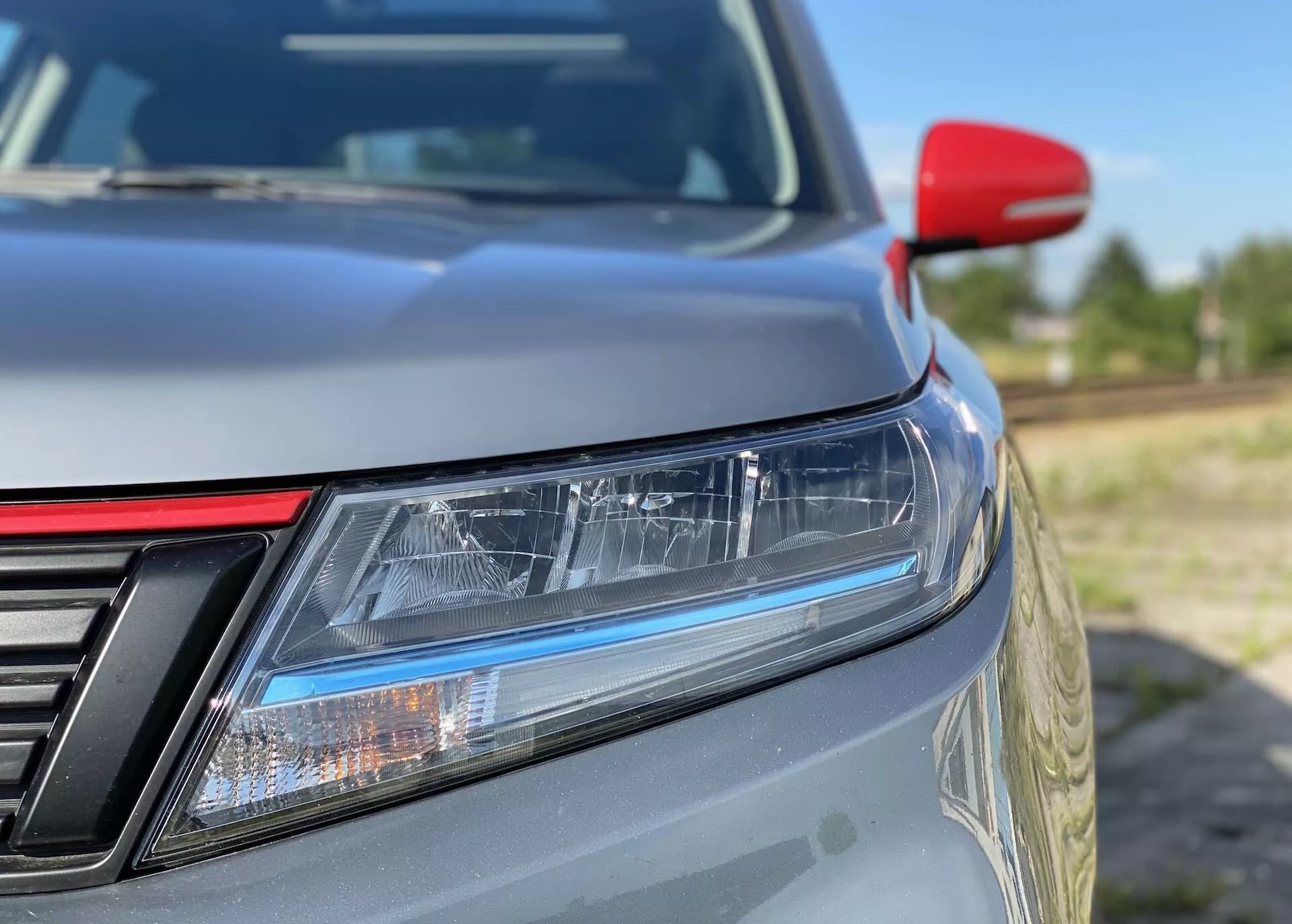 Toma de primer plano de la esquina frontal izquierda de un Suzuki Vitara 2018, enfocándose en características detalladas como faros LED con luces diurnas, una parrilla parcialmente visible con barras verticales, el capó con una suave inclinación, pintura metálica, un espejo lateral rojo contrastante posiblemente con indicadores de giro integrados y un fondo desenfocado que resalta el diseño elegante y la eficiencia aerodinámica.