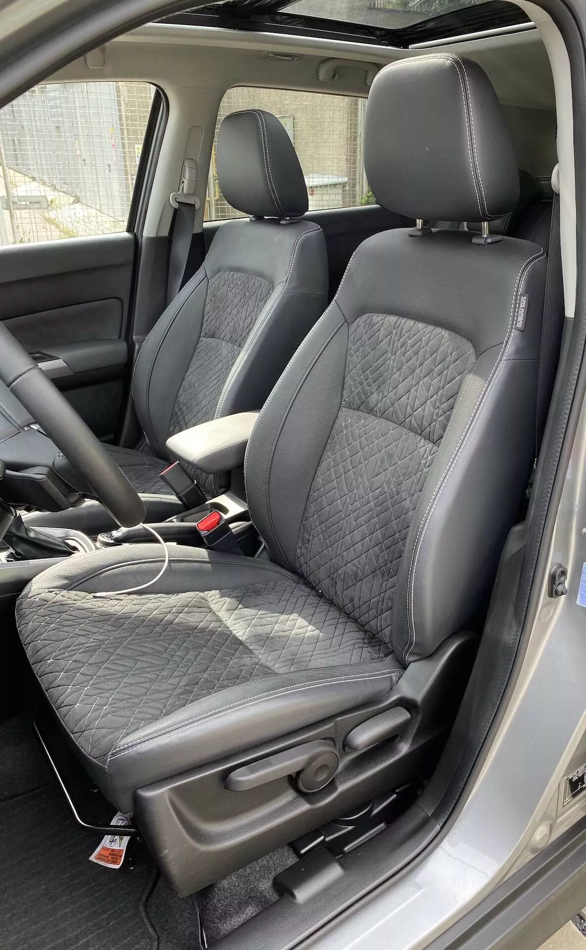 El interior de un Suzuki Vitara 2018, destacando los asientos delanteros estilo cubo moderno desde una perspectiva ligeramente sobre el nivel del asiento. Los asientos están tapizados en cuero negro con un inserto acolchado en patrón de diamantes y refuerzos laterales de cuero liso, ofreciendo un equilibrio entre comodidad y deportividad. Una etiqueta roja en los asientos sugiere características adicionales como airbags laterales o asientos calefactados. Es visible la consola central con un reposabrazos cubierto de cuero, así como un cable de carga en el asiento del pasajero, lo que indica la presencia de puertos de carga. Los hebillas de los cinturones de seguridad combinan con el esquema de color negro del interior. También es visible parte del tablero frontal y el pilar B, sugiriendo una cabina espaciosa.