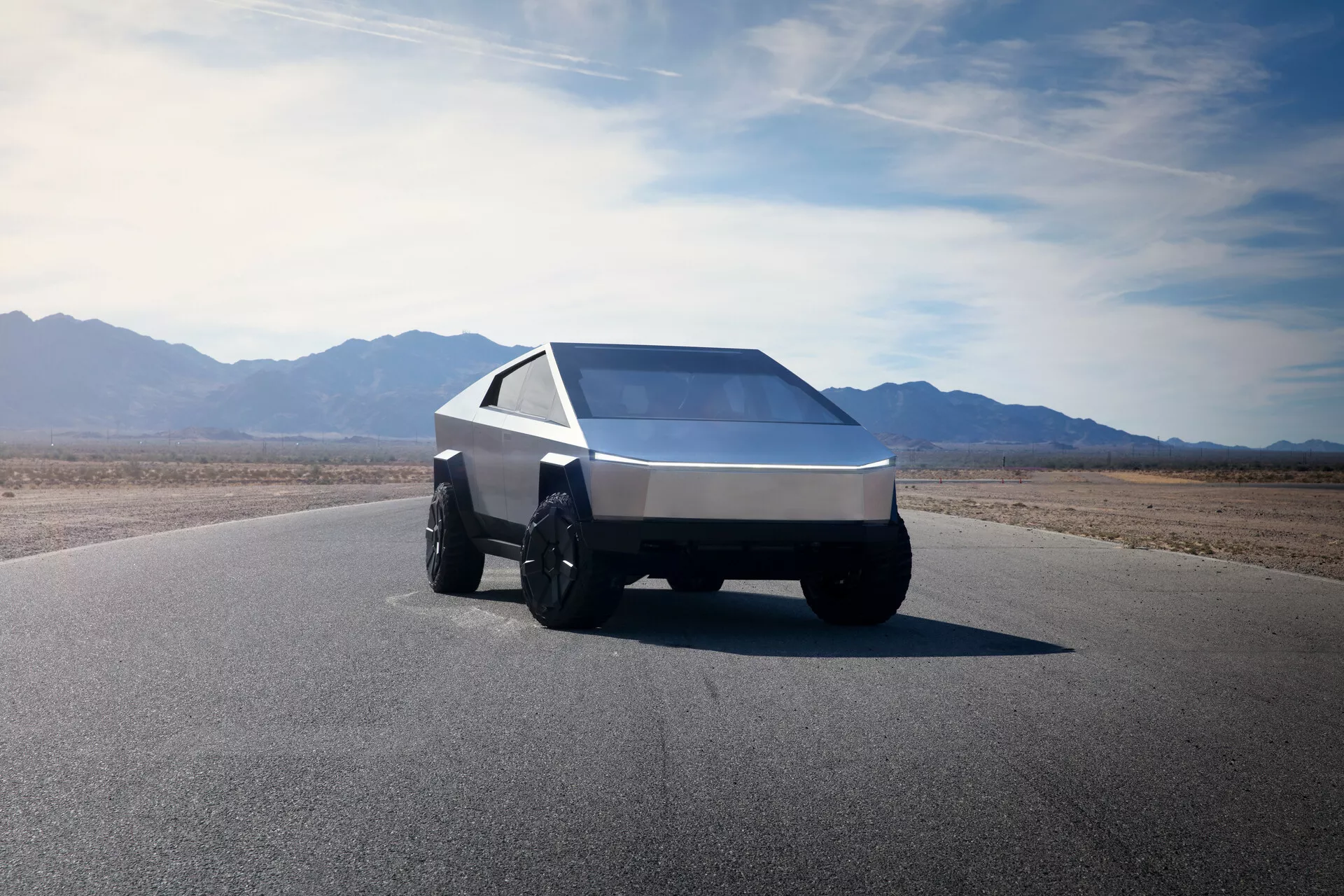 Un Tesla Cybertruck 2021 (1ª generación) fotografiado desde un ángulo bajo de tres cuartos delantero, mostrando su diseño angular y futurista. Esta vista captura el lado delantero izquierdo del coche, enfatizando su forma en cuña con líneas afiladas y superficies planas. El vehículo está equipado con grandes y resistentes neumáticos adecuados para la conducción todoterreno y presenta una única tira de luz continua a lo largo del ancho del frente, indicativa de la moderna iluminación LED o láser. El parabrisas inclinado y expansivo y las ventanas laterales afiladas contribuyen a su estética geométrica. El coche está hecho de un metal cepillado o material compuesto, sugiriendo durabilidad y robustez. El fondo incluye un cielo claro y expansivo y un terreno montañoso, insinuando un entorno árido y desértico.