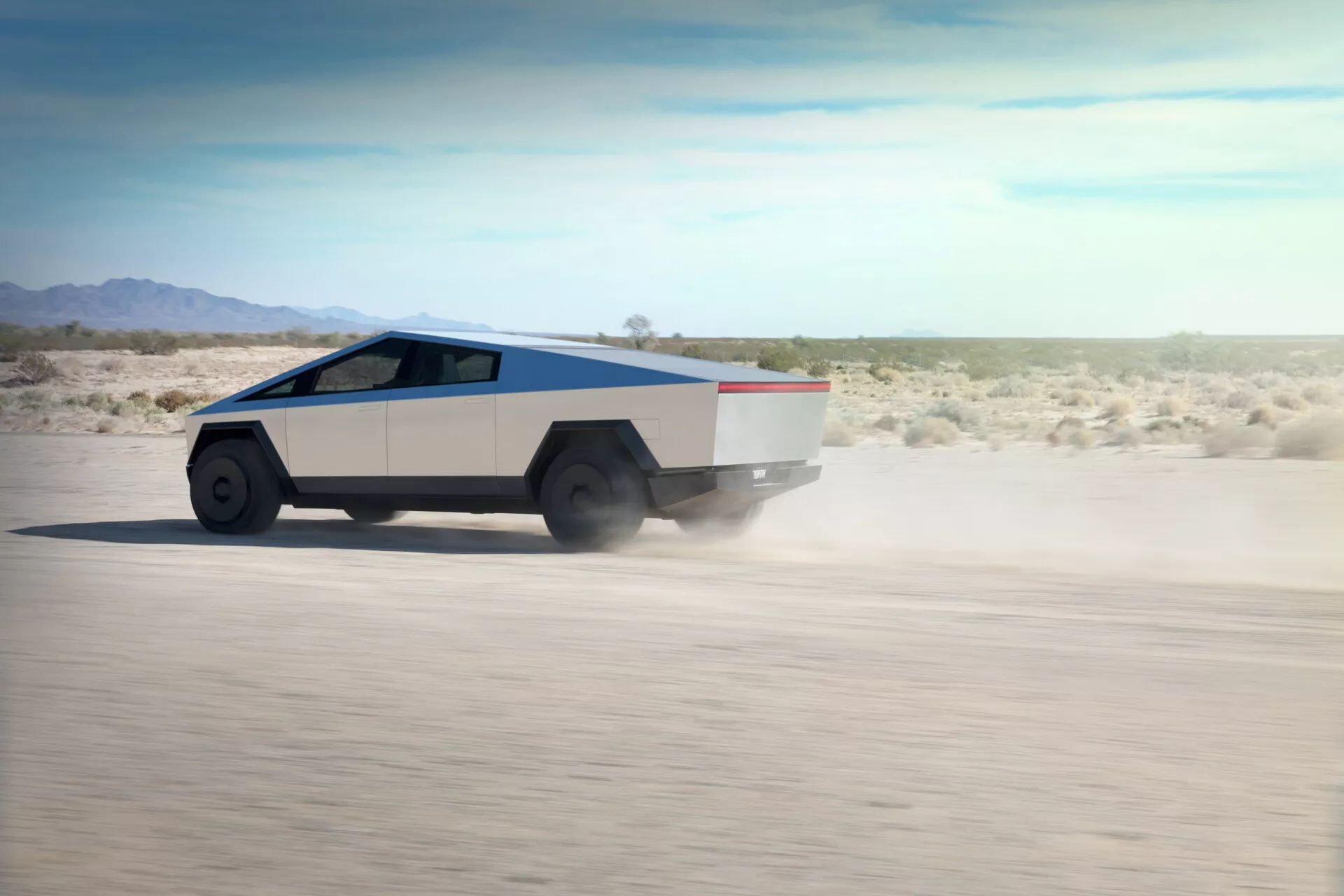 Esta imagen muestra un Tesla Cybertruck 2021 en movimiento sobre un terreno desértico plano, visto desde un ángulo trasero de tres cuartos que resalta su lado derecho y trasero. La camioneta tiene un diseño geométrico y angulado con un perfil en forma de cuña, una carrocería de dos tonos, plateada y gris claro, con una franja contrastante a lo largo de la línea de cintura, y un techo inclinado. Las ruedas grandes y robustas con neumáticos todoterreno complementan el diseño angular. El polvo levantado indica alta velocidad. Las ventanas laterales parecen fusionarse en una pieza continua de vidrio desde el parabrisas hasta la parte trasera. Probablemente un vehículo eléctrico dado el diseño avanzado y la ausencia de tubos de escape.