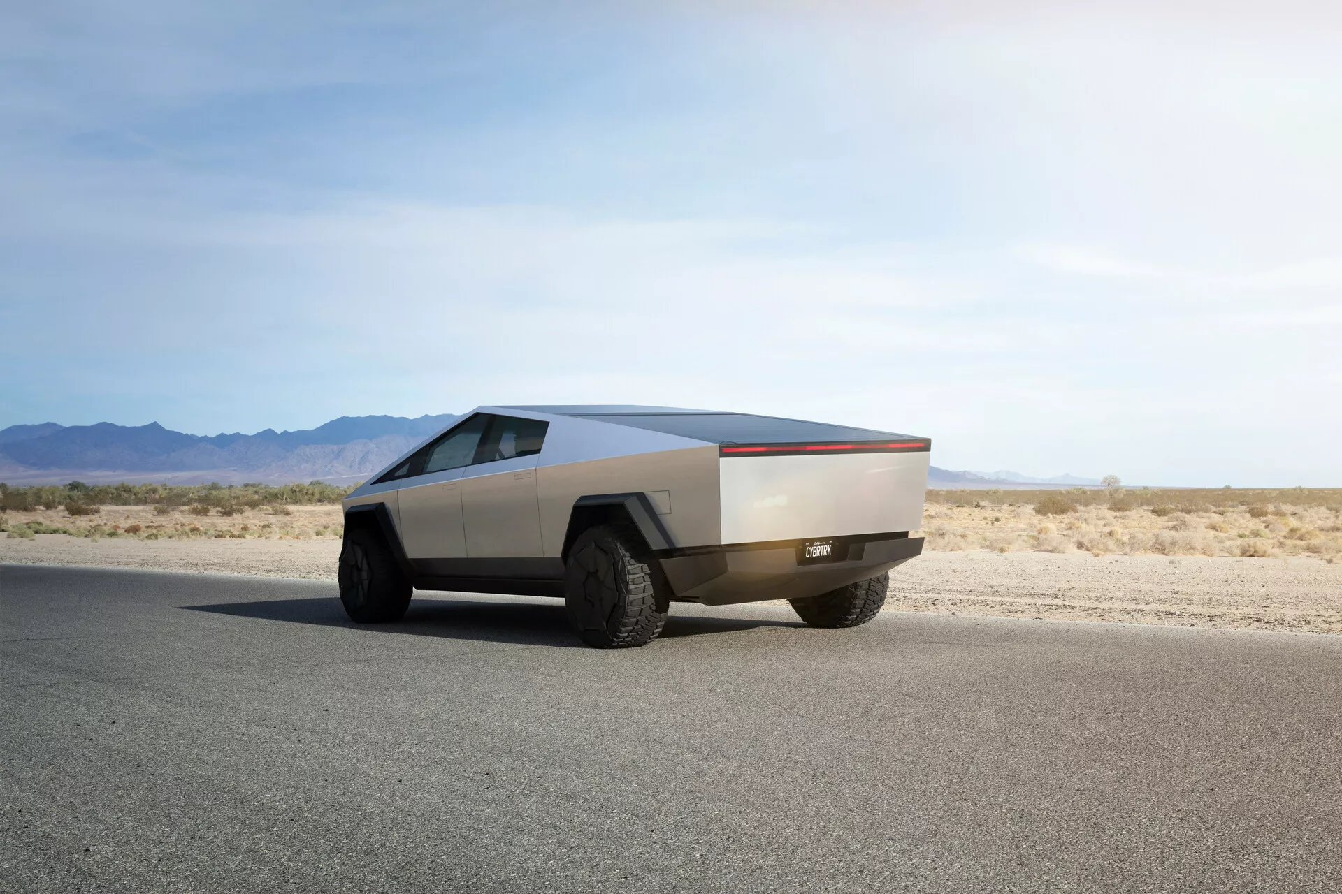 Futurista Tesla Cybertruck 2021 en carretera asfaltada plana capturada desde un ángulo de tres cuartos trasero, destacando su diseño angular distintivo, acabado metálico, grandes neumáticos todoterreno y ausencia de espejos y manijas de puertas tradicionales, lo que indica mecanismos automáticos avanzados.