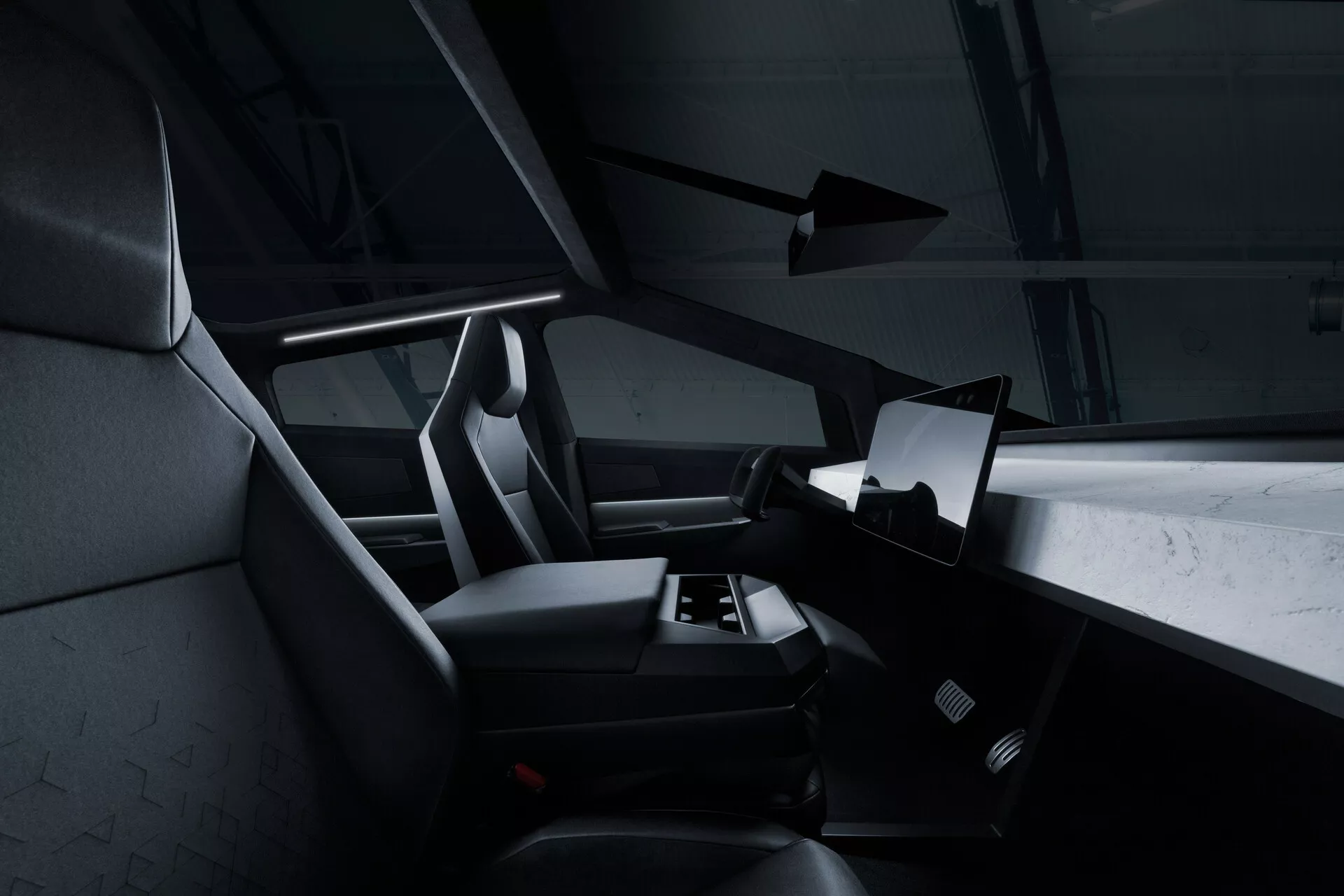 Vista interior del Tesla Cybertruck 2021 (1ª Generación) desde la perspectiva del asiento trasero, mostrando el lado derecho de la cabina con una gran pantalla táctil horizontal dominando el centro del tablero. Parte del volante es visible, pero mayormente oculto. Los asientos del pasajero delantero y del conductor tienen reposacabezas integrados y tapicería texturizada, posiblemente de tela o cuero sintético. Se vislumbra una alfombrilla en el área de los pies y no se ven controles manuales, lo que sugiere un enfoque minimalista y digital. La paleta de colores oscuros y la sutil iluminación de acento dan una sensación moderna y lujosa.