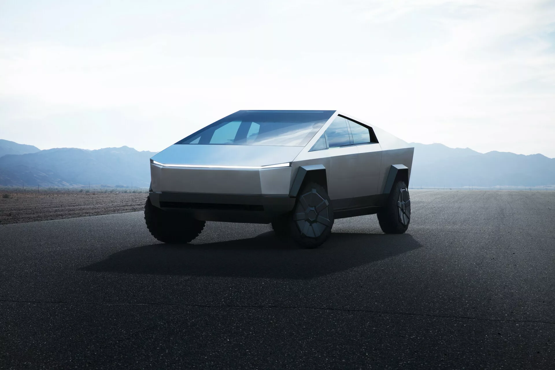 Imagen del Tesla Cybertruck 2021 (1ª generación) en una carretera con terrenos planos o montañas desérticas en el fondo, durante el día con cielos despejados. La foto está tomada desde un ángulo bajo, proporcionando una vista de cuarto delantero que resalta el diseño angular y futurista del camión. El vehículo presenta un frente en forma de cuña, cuerpo de paneles planos, faros horizontales delgados, un parabrisas de pendiente continua, sin espejos laterales visibles, grandes ruedas aptas para todo terreno con llantas intrincadas y carece de una parrilla frontal, indicando su propulsión eléctrica.