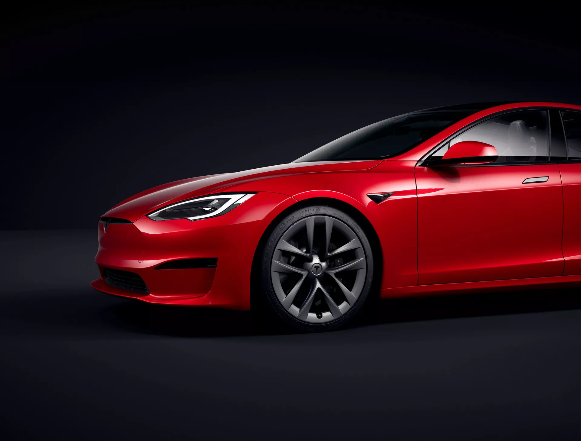 Tesla Model S 2016 (facelift de 1ra generación) sedán rojo en estudio con iluminación controlada y fondo negro, vista desde un ángulo bajo que enfatiza el perfil aerodinámico y la holgura del suelo, vista frontal tres cuartos mostrando el lado del conductor, faros, rejilla, parte del capó, diseño de llantas de aleación premium con patrón de múltiples radios, parabrisas con vidrio tintado, línea de techo inclinada, espejo lateral, manijas de puerta y línea de ventana integrándose suavemente en el diseño elegante, resaltando el acabado de la pintura, líneas de contorno y paneles esculpidos sugiriendo lujo y desempeño, iluminación enfocada mostrando detalles de diseño.