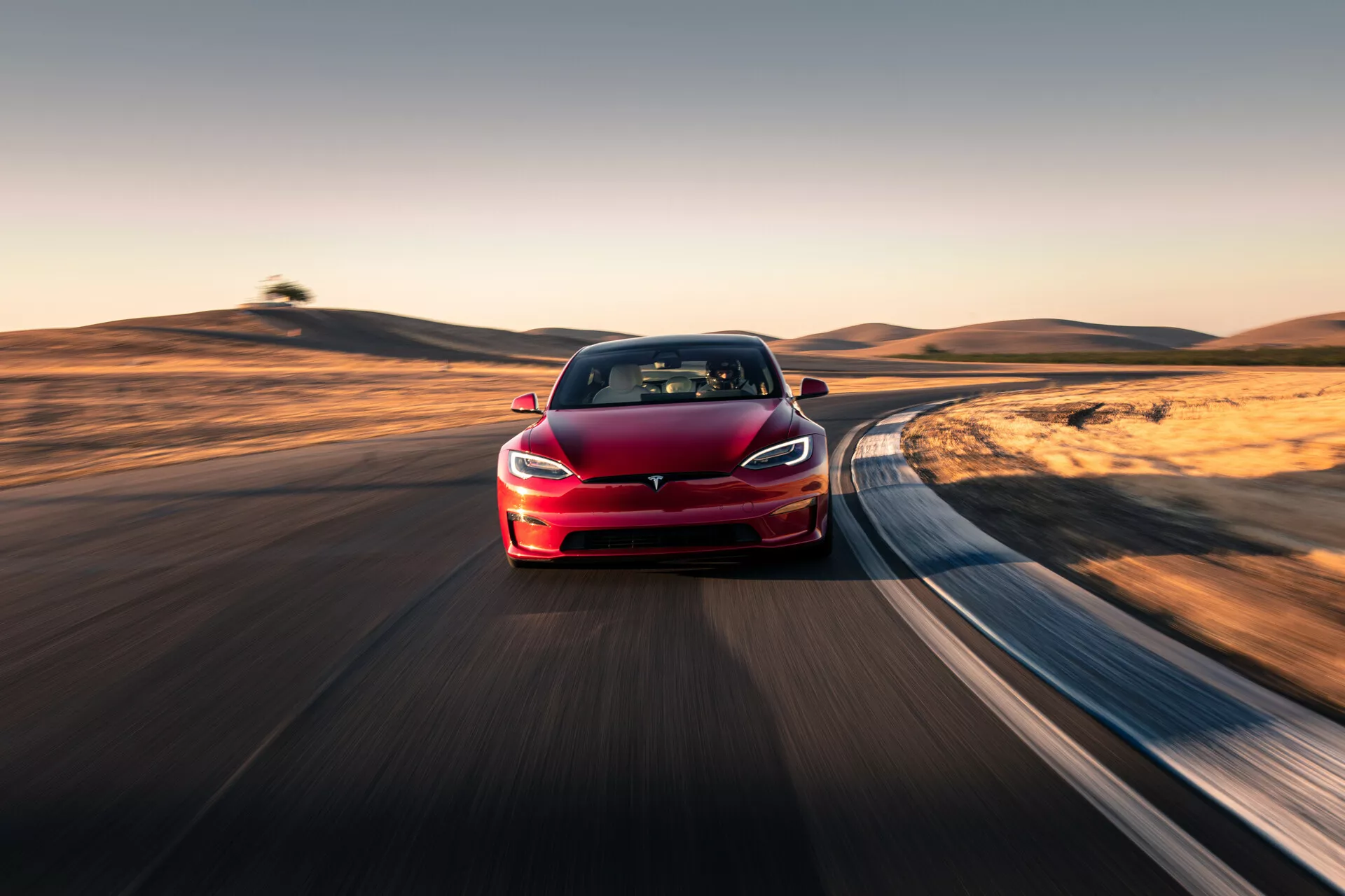 Imagen de un sedán rojo Tesla Model S 2016 (primera generación con renovación) conduciendo en una carretera curva, capturada desde un ángulo de tres cuartos frontal. La foto enfatiza el lado izquierdo delantero del vehículo, mostrando el emblema, los faros y parte del capó, extendiéndose hasta la parte trasera. La ausencia de una parrilla tradicional resalta su naturaleza eléctrica. El fondo está desenfocado con un efecto de movimiento, indicando velocidad y posiblemente logrado mediante la técnica de paneo. El auto, posicionado cerca de la cámara con una perspectiva comprimida, refleja avances tecnológicos modernos como un sistema de propulsión avanzado, probablemente piloto automático/sistemas de asistencia al conductor y luces LED. La escena bien iluminada durante el día con sombras largas sugiere un entorno de madrugada o tarde, con un paisaje natural que ofrece un contraste dinámico con el diseño moderno del vehículo.