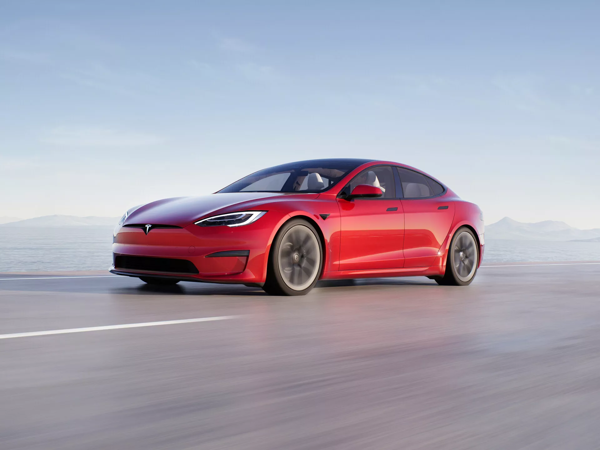 La imagen muestra un sedán moderno de cuatro puertas Tesla Model S 2016 (facelift de la primera generación) en movimiento sobre una carretera o pista lisa. Presentado desde un perfil lateral a baja altura, las líneas elegantes y aerodinámicas del vehículo son claramente visibles, destacando su distintiva parrilla cerrada, faros LED delgados y parachoques delantero suave. El diseño fastback con una línea de techo suavemente inclinada, manijas de puertas empotradas retráctiles y espejos retrovisores laterales minimalistas resaltan el enfoque en la aerodinámica del coche. Las grandes llantas de múltiples radios sugieren una optimización para el flujo de aire y la reducción de la resistencia. La composición utiliza desenfoque de movimiento para transmitir alta velocidad, con el coche en sí mismo nítidamente enfocado contra un fondo borroso. Esta fotografía profesional enfatiza la naturaleza eléctrica del coche, su iluminación avanzada, diseño aerodinámico y posibles características de asistencia al conductor.