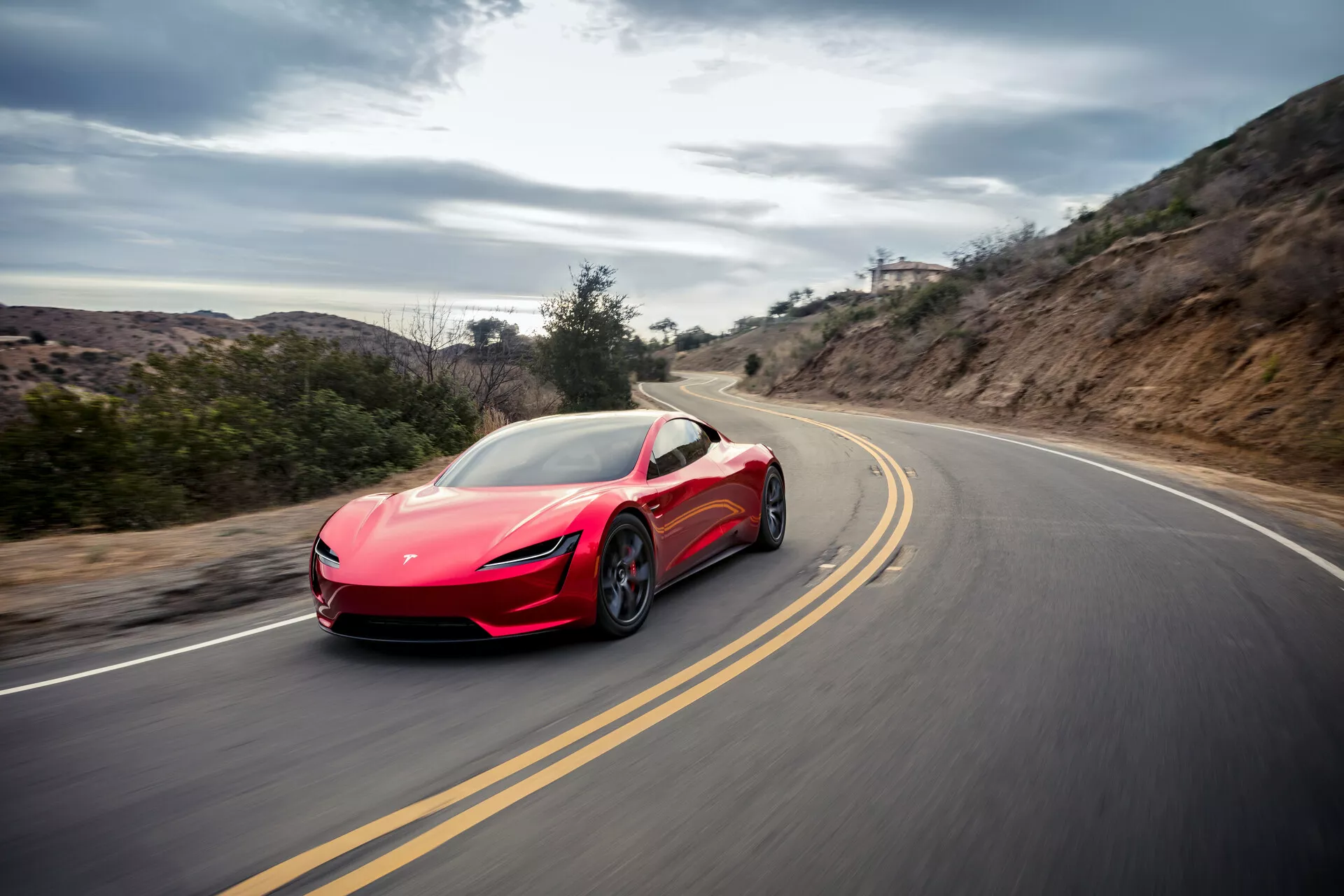 Toma dinámica de un coche deportivo Tesla Roadster 2020 (segunda generación) de color rojo capturado en movimiento en una carretera sinuosa que muestra sus líneas aerodinámicas, faros elegantes, espejos laterales, grandes ruedas y tomas de aire, contra un fondo de luz difusa que indica una conducción a alta velocidad en un entorno rural.