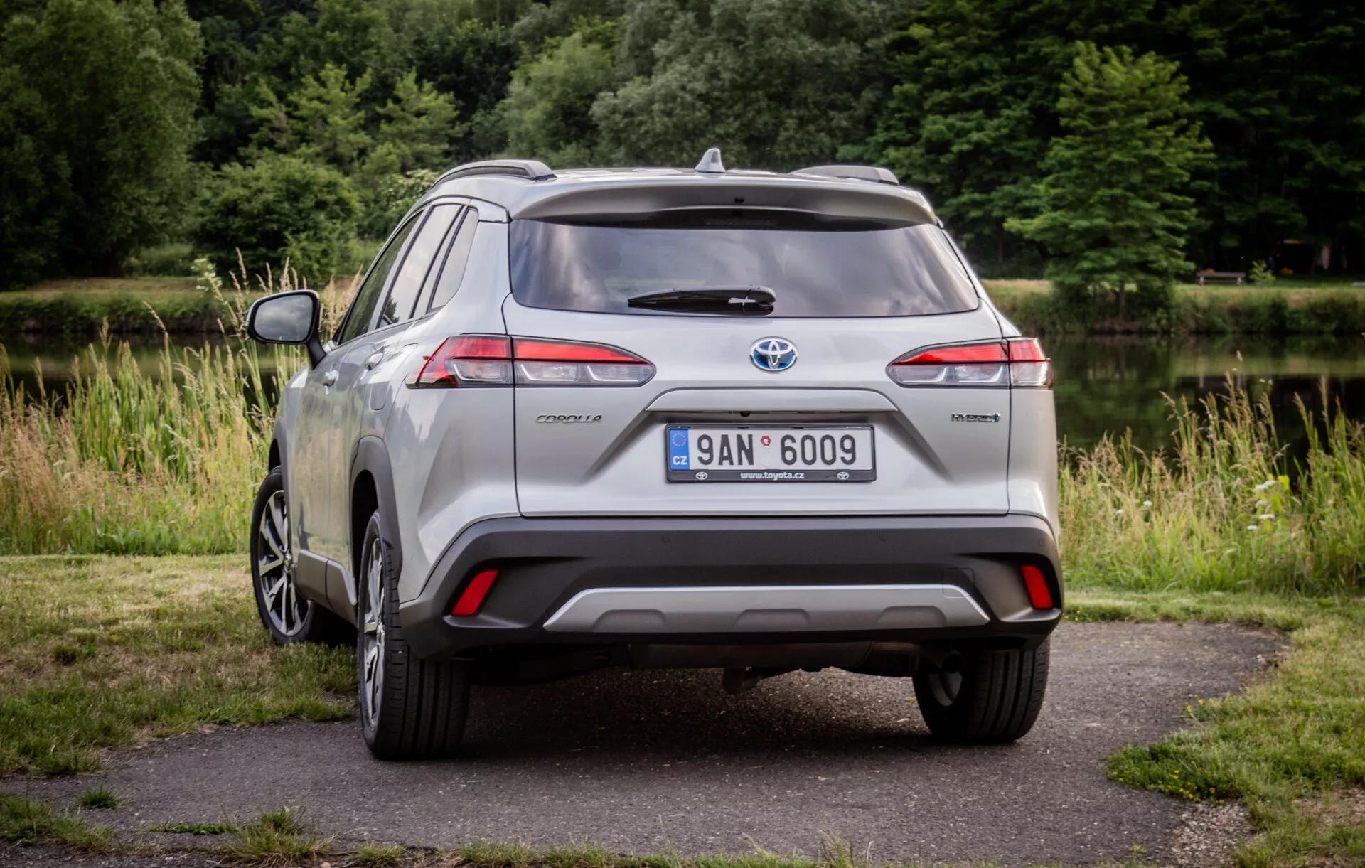 Toyota Corolla Cross plateado del 2022 estacionado sobre pavimento con vegetación al fondo, mostrando sus luces traseras LED, spoiler trasero, rueda de aleación de múltiples radios y distintivo híbrido a la luz del día