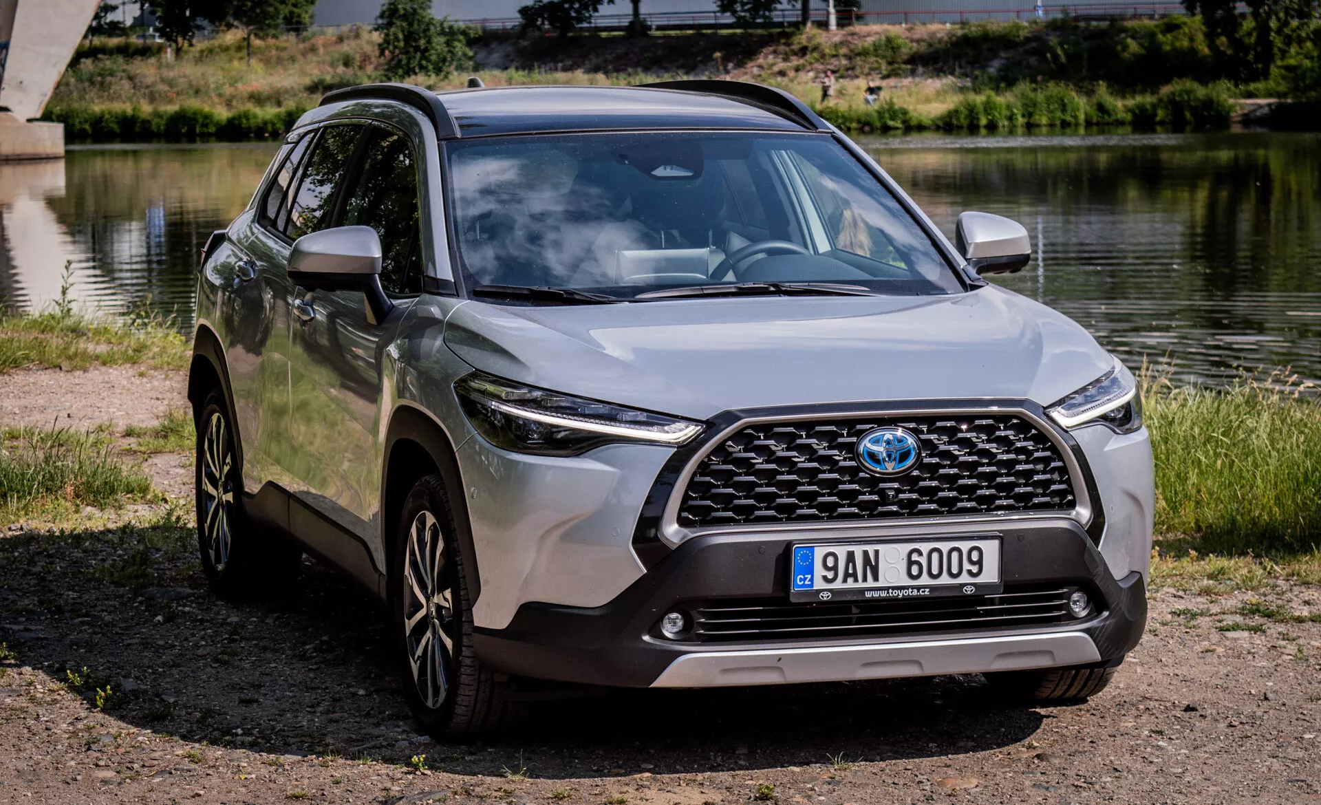 SUV crossover moderno Toyota Corolla Cross 2022 estacionado al aire libre cerca de un cuerpo de agua con vista clara de su parrilla frontal, capó, faros LED, parachoques frontal y perfil lateral que incluye ruedas de aleación y espejos laterales del color de la carrocería