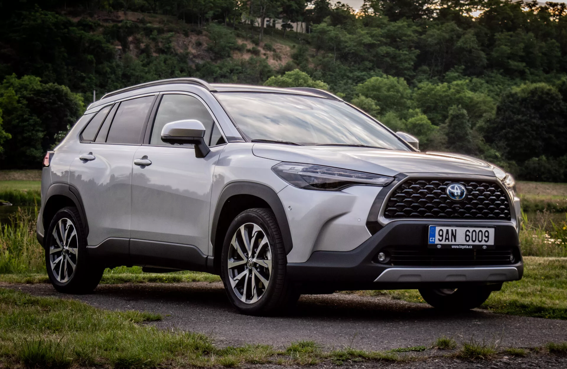 Toyota Corolla Cross 2022 con acabado metálico brillante que muestra su diseño moderno de SUV con parrilla frontal detallada, emblema, faros LED angulares, parachoques frontal aerodinámico, capó contorneado, perfil lateral con ruedas de aleación de múltiples radios y barras en el techo, tomada desde un ángulo bajo para enfatizar su altura y postura durante el día en un entorno suburbano natural.
