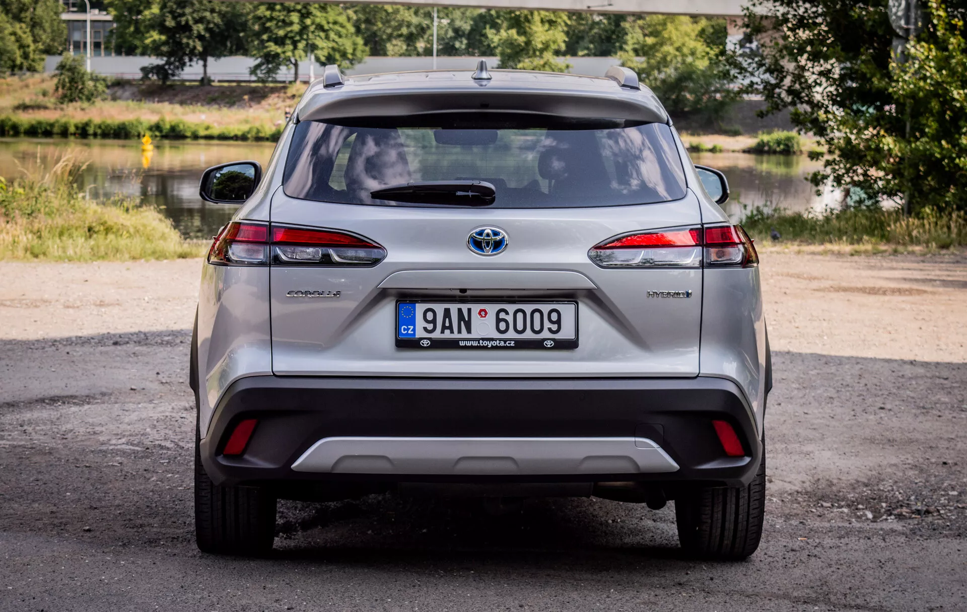 Vista trasera del Toyota Corolla Cross 2022, mostrando luces traseras LED, insignia híbrida y emblema de Toyota cromado con luz natural diurna y un escenario pintoresco de vegetación y agua