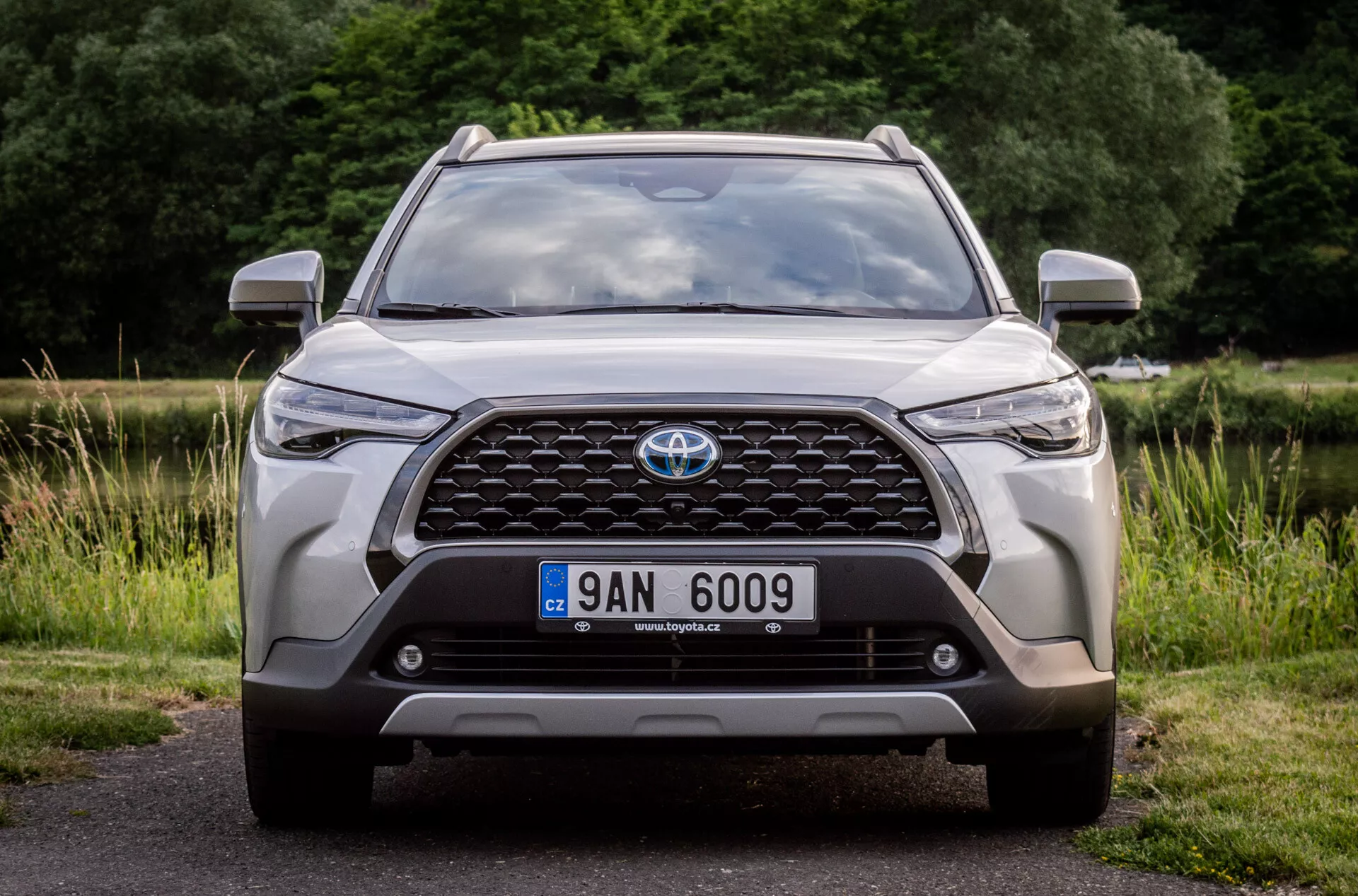 Vista frontal de un Toyota Corolla Cross 2022, destacando su amplia parrilla con emblema, faros modernos LED, luces antiniebla, posición de la placa de matrícula, sensores de estacionamiento integrados o cámara frontal, contornos aerodinámicos del capó, diseño rugoso del parachoques inferior y tomas de aire, estacionado en asfalto con un fondo natural
