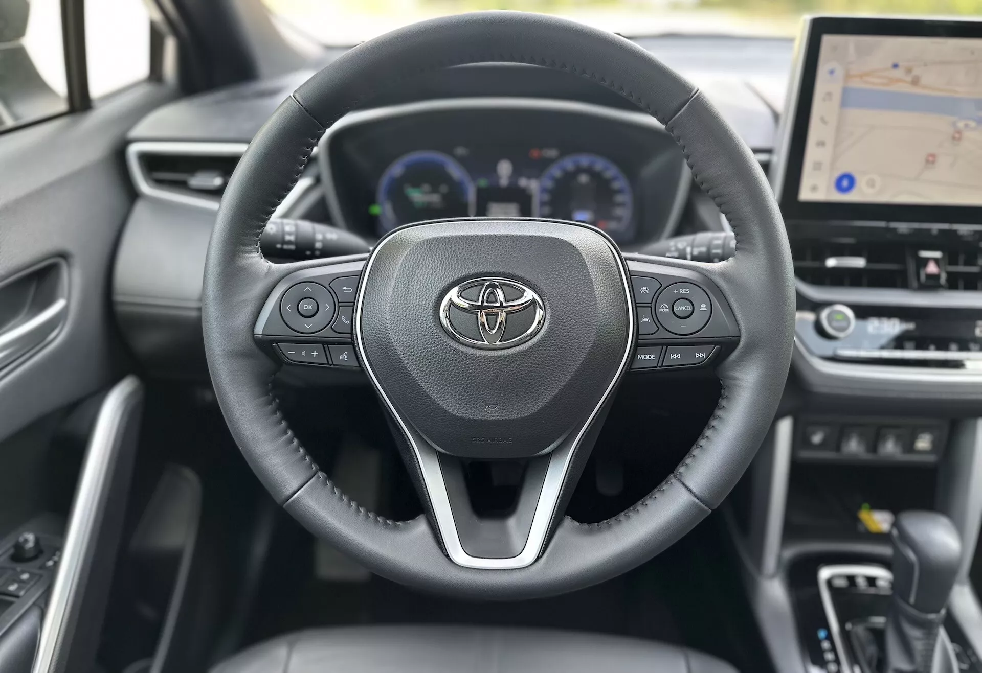 Interior del Toyota Corolla Cross 2022 mostrando el volante con el logotipo de Toyota, controles integrados para audio, llamadas y control de crucero, detrás de los cuales se ve parcialmente un tablero digital con velocímetro, tacómetro y pantalla multifuncional, incluido un display central más grande para la navegación GPS, todo ambientado contra un diseño de material oscuro contemporáneo.