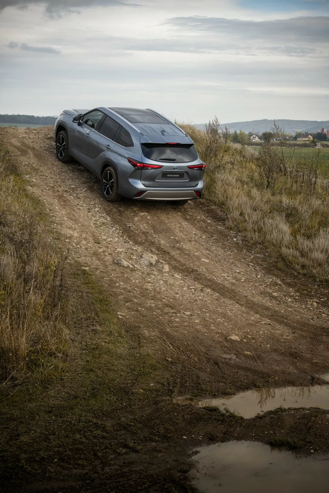 Toyota Highlander oscuro 2021 capturado desde un ángulo alto en un camino de tierra, mostrando la vista trasera de tres cuartos, diseño deportivo y potencial para viajes ligeros todo terreno entre un paisaje campestre