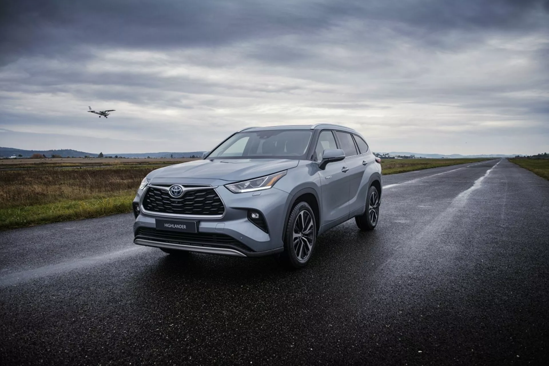 SUV crossover Toyota Highlander 2021 en carretera de asfalto mojado, mostrando sus faros angulares, parrilla frontal prominente, ruedas de aleación de múltiples radios y diseño aerodinámico bajo iluminación difusa y nublada