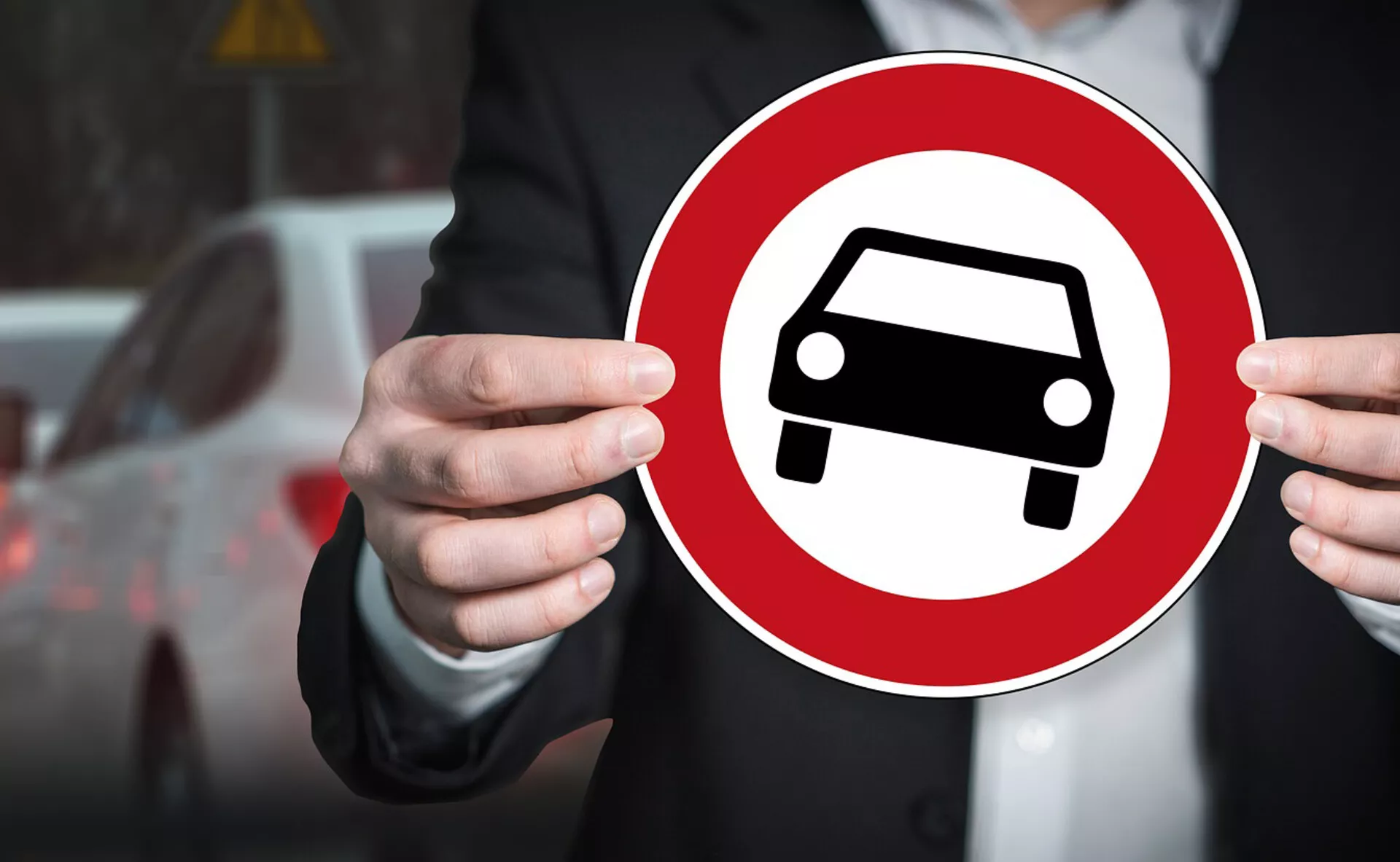 Individuo sosteniendo un letrero circular rojo con un icono de silueta de coche blanco, simbolizando la representación genérica de un automóvil contra un fondo urbano borroso