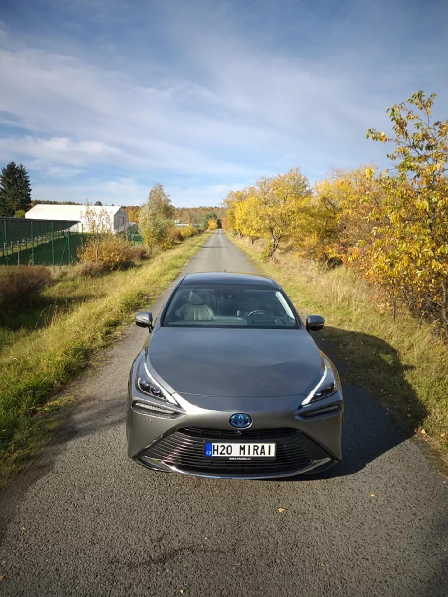 Toyota Mirai 2021 fotografiado desde un ángulo frontal y alto, destacando sus elegantes faros LED y la prominente parrilla con emblema central que posiblemente incluye tecnología de sensores, en un contexto semi-rural bajo luz natural, mostrando avanzada tecnología de celda de combustible de hidrógeno y diseño aerodinámico