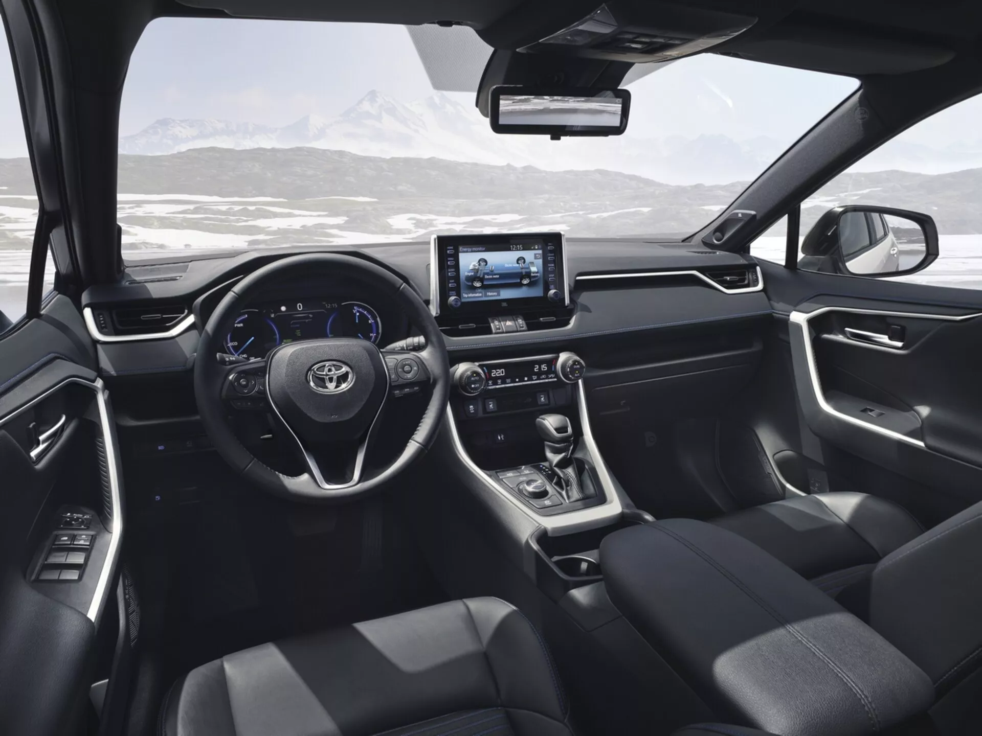Vista interior del Toyota RAV4 2019 desde el asiento trasero, mostrando el diseño moderno y las características tecnológicas incluyendo el volante con botones de control, pantalla digital, sistema de infotenimiento táctil, palanca de cambios de transmisión automática, asientos tapizados en oscuro, y controles en el panel de la puerta con un paisaje montañoso nevado visible a través del parabrisas.