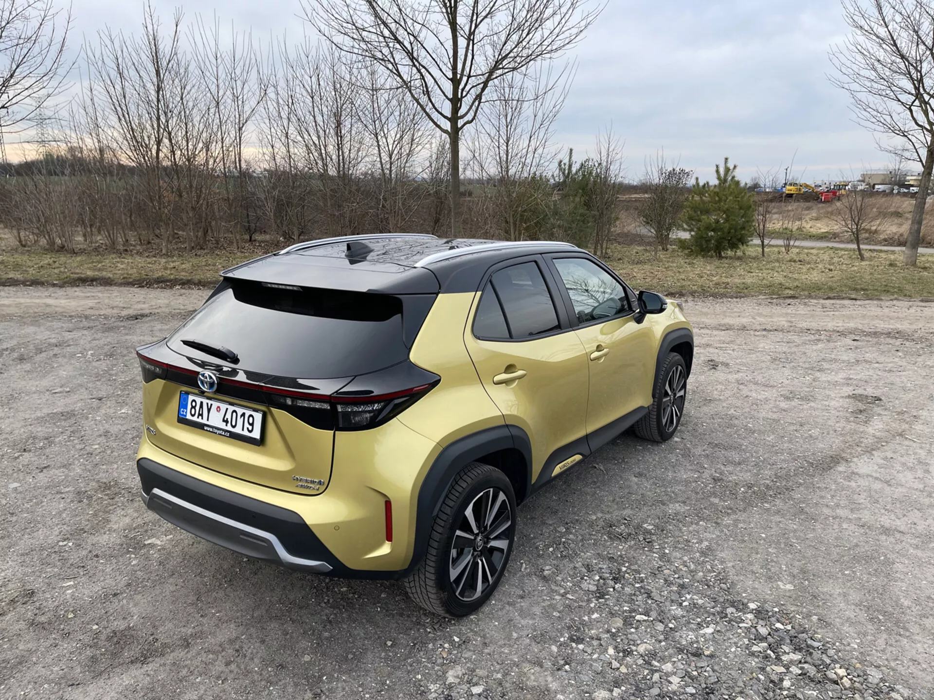 Toyota Yaris Cross 2021 capturado desde una vista trasera de tres cuartos destacando su spoiler aerodinámico, pintura metálica dorada y luces traseras LED, estacionado en una superficie sin pavimentar con fondos naturales e industriales.