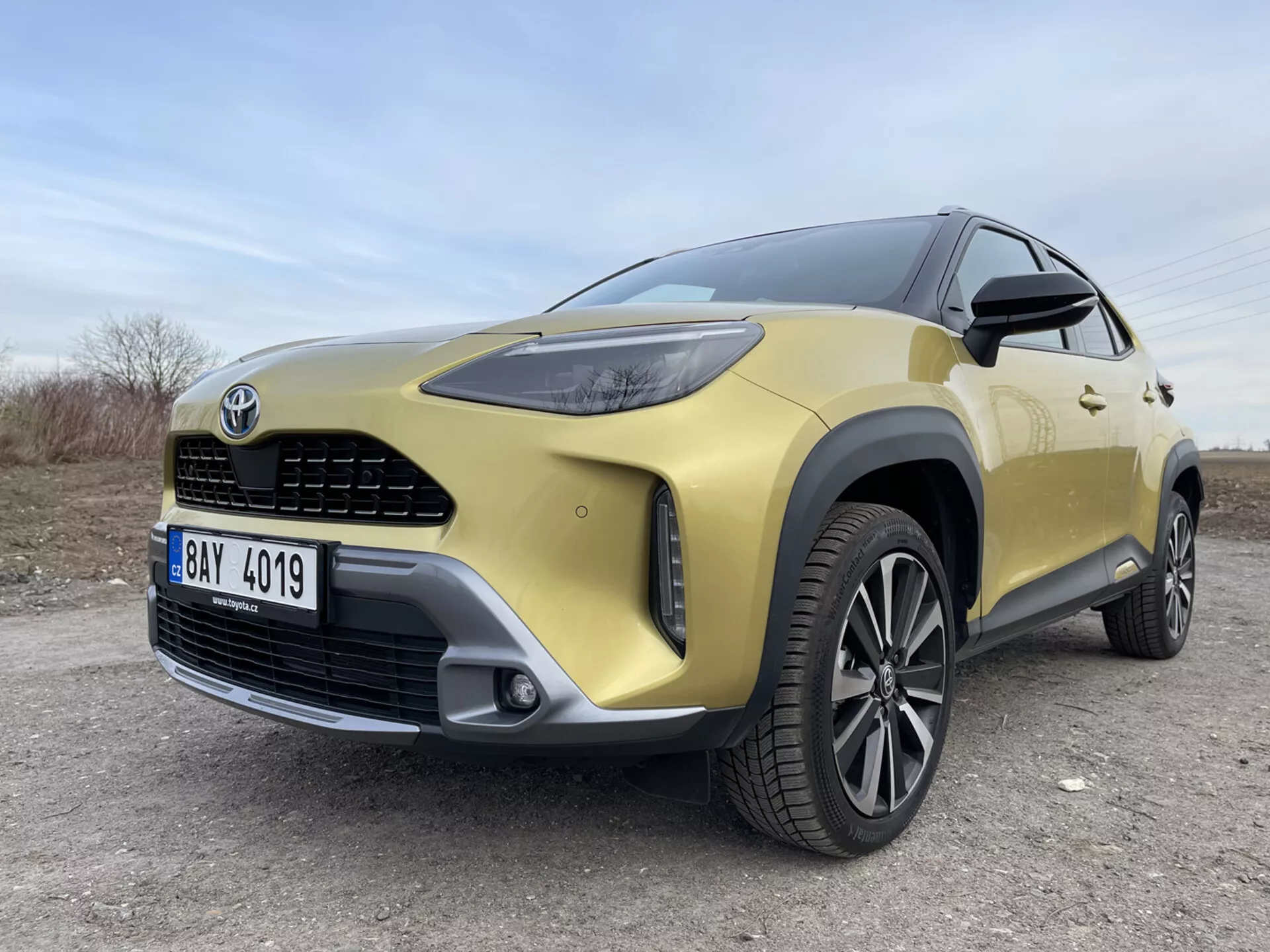 SUV Toyota Yaris-Cross 2021 en oro mostrando su cuarto delantero izquierdo desde una vista angulada y baja resaltando los faros LED, la parrilla frontal, el parachoques, la llanta de aleación y elementos de diseño moderno sobre una superficie no pavimentada