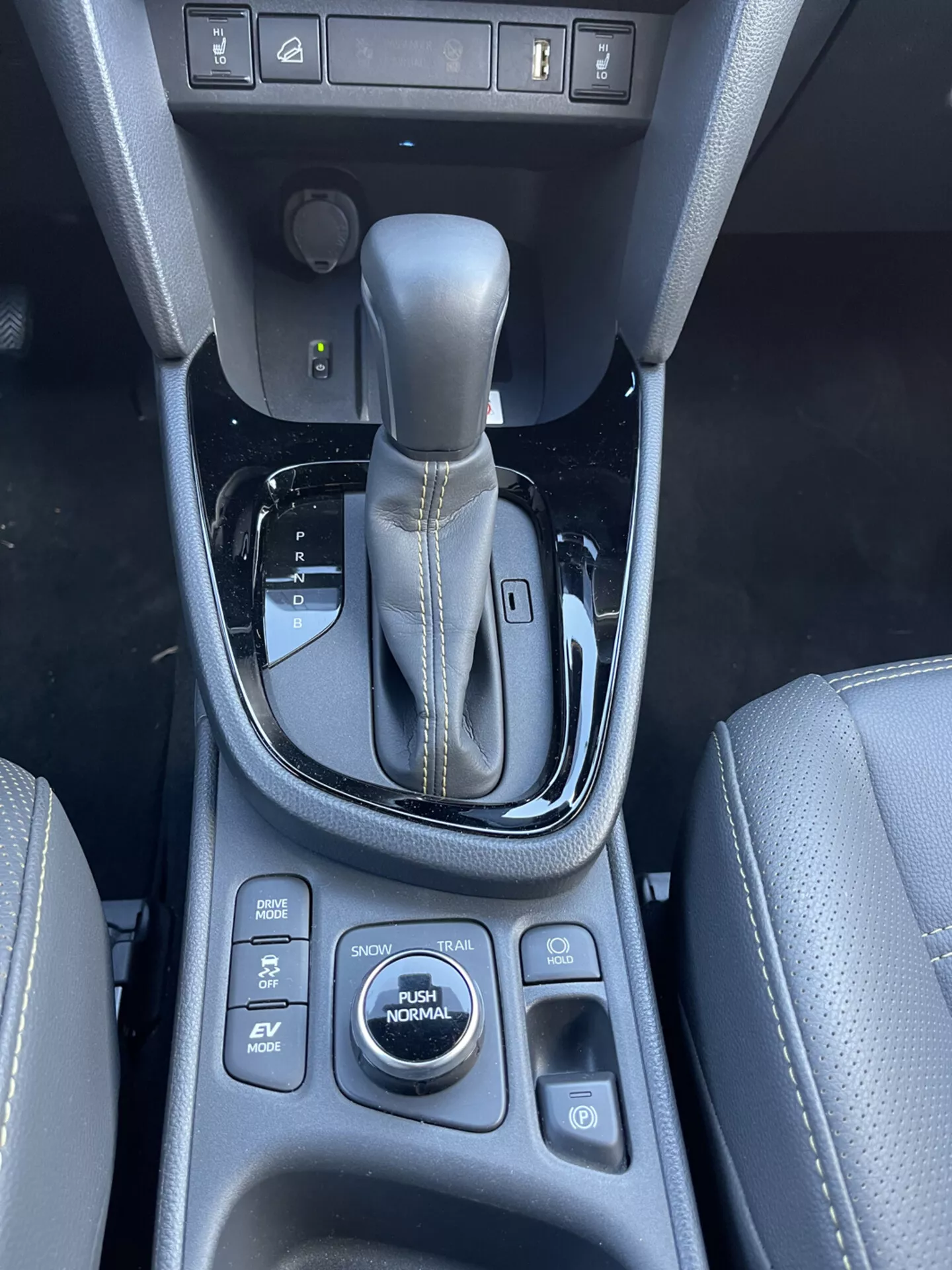 Interior del Toyota Yaris-Cross 2021 que muestra la consola central, palanca de cambios automática en posición de estacionamiento con ribete metálico y bisel negro brillante, botones de selección de modo de conducción incluyendo SPORT, ECO, EV, NORMAL, control de tracción, controles de calefacción de asientos y un enchufe de 12V, enfatizando la calidad del diseño y las características avanzadas de la tecnología del vehículo.