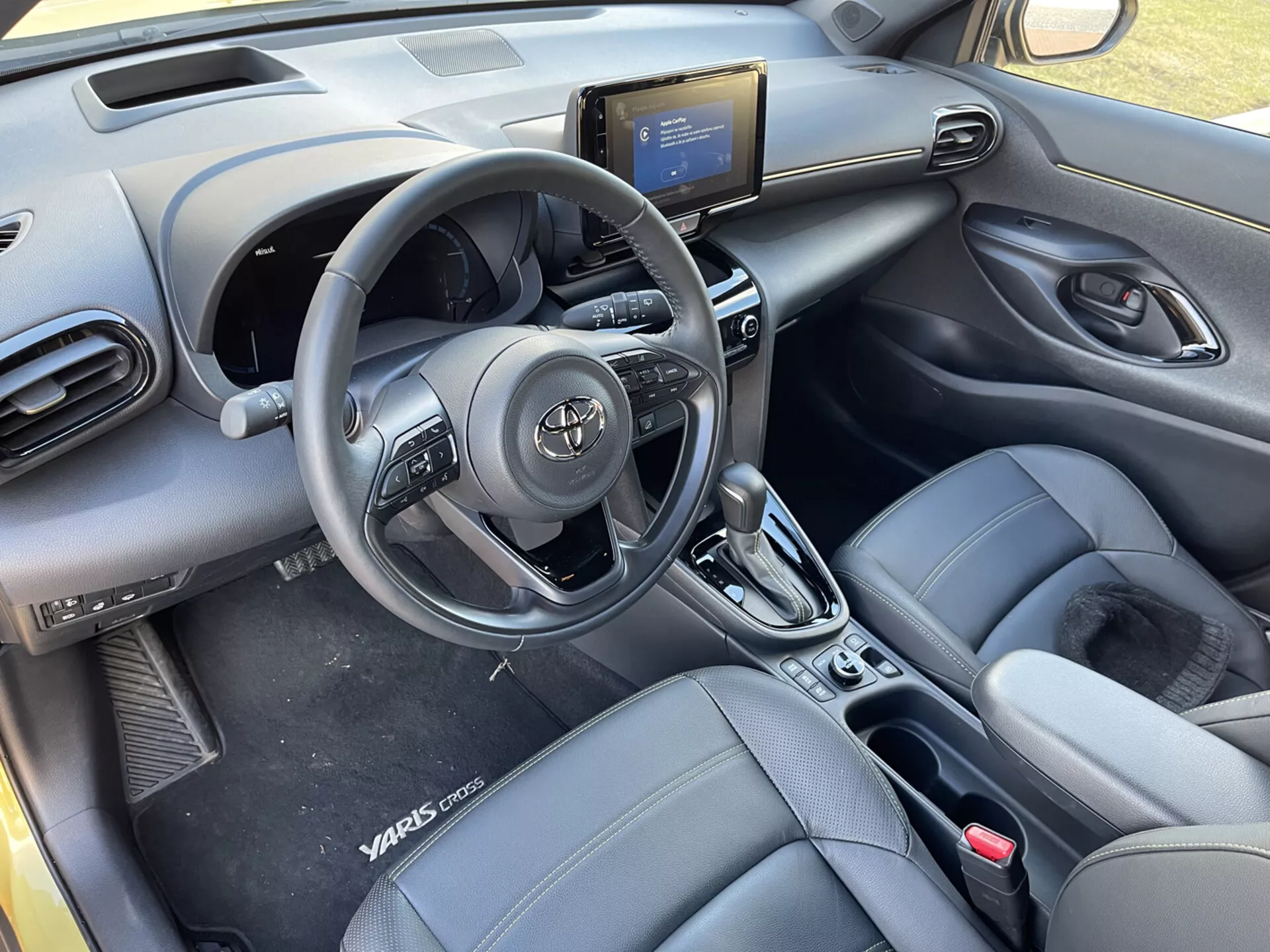 Vista interior del Toyota Yaris-Cross 2021 desde los asientos traseros, destacando volante con controles, salidas de aire en el tablero, consola central con sistema de infoentretenimiento, controles de aire acondicionado, palanca de transmisión automática, asientos delanteros tipo cubo con aspecto de cuero con marca Yaris Cross, paneles de puerta con controles eléctricos, y un diseño de cabina moderno y elegante.