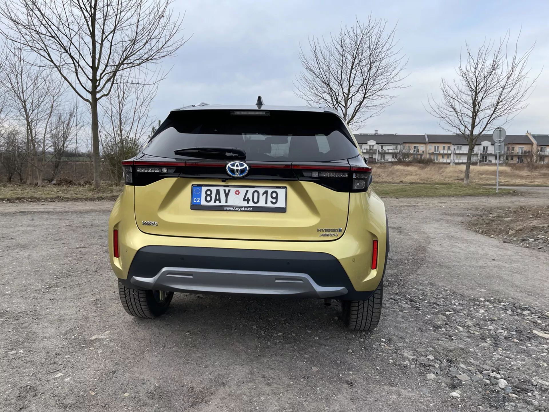 Vista trasera de un Toyota Yaris Cross amarillo 2021 capturada a nivel de los ojos mostrando su diseño moderno, conjunto de luces traseras horizontal, emblema de Toyota, nombre del modelo Urban Cruiser, placa de registro, parachoques trasero de diseño contrastante con sensores de estacionamiento integrados y manija del maletero, ambientado en un fondo suburbano.