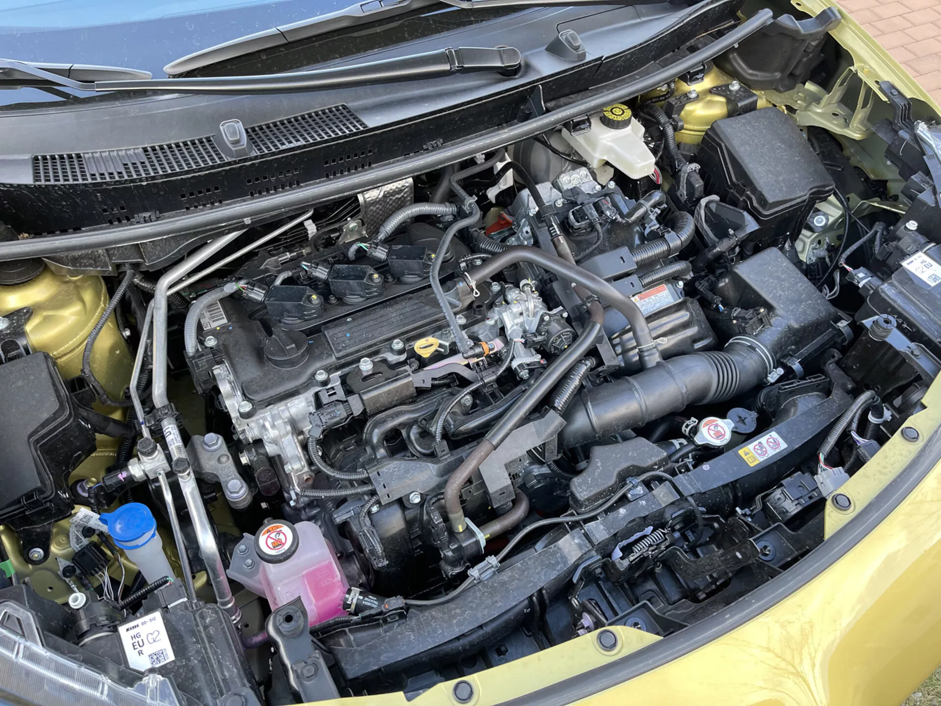 Vista detallada del compartimiento del motor del Toyota Yaris-Cross 2021 mostrando un bloque de motor en línea, cubierta de motor de plástico negro, sistema de admisión de aire, depósitos de refrigerante y líquido de frenos, componentes eléctricos, batería, radiador y un diseño ordenado con etiquetas de seguridad.