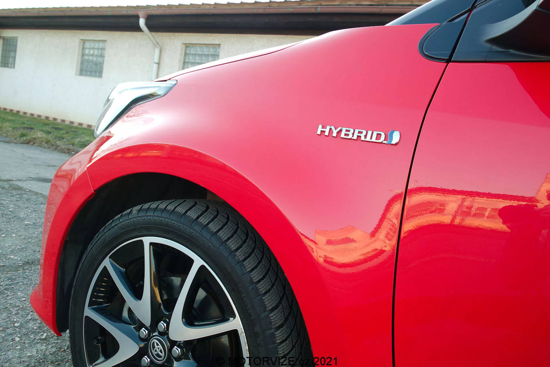 Vista en primer plano de un Toyota Yaris 2020 rojo, centrado en el panel del cuarto frontal y la rueda de aleación de varios radios. La insignia híbrida es visible, destacando la tecnología ecológica del vehículo. La pintura brillante refleja la luz del sol, resaltando el acabado de alta calidad y el diseño aerodinámico y moderno del coche.