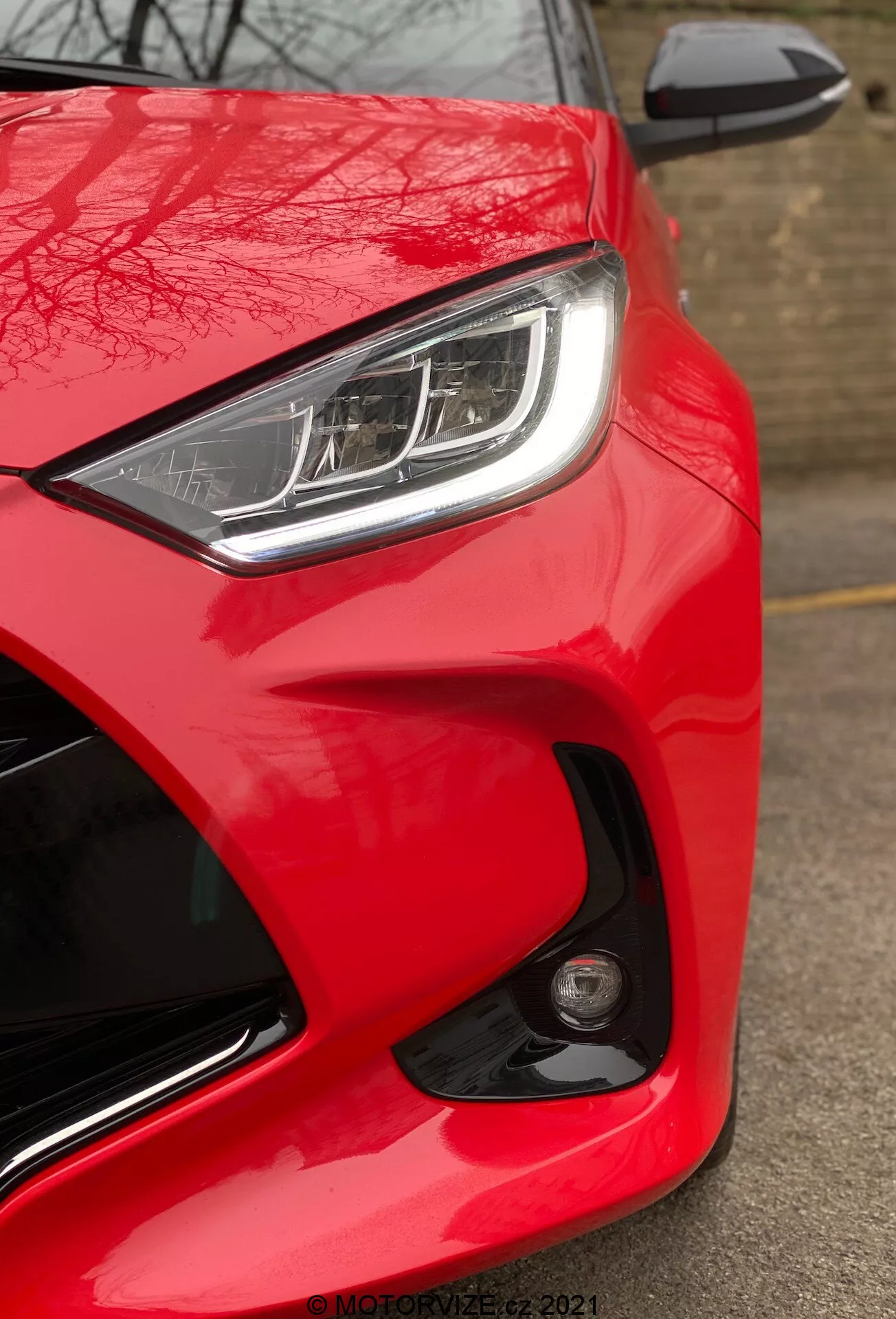 Vista de cerca del Toyota Yaris 2020, enfocado en la parte delantera del lado del conductor desde un ángulo bajo. La imagen resalta el color rojo brillante y brillante del automóvil, el faro LED o HID elegante con luces diurnas distintivas y el espejo lateral del color de la carrocería parcialmente visible con indicador de señal de giro integrado. El diseño aerodinámico se enfatiza a través de las líneas y curvas esculpidas alrededor del faro y el capó. Un faro antiniebla redondo o sensor en el paragolpes delantero sugiere características de seguridad avanzadas. La falta de desenfoque por movimiento indica que el automóvil está detenido, y se ve una marca de agua 'MOTORZETZ. 2021' en la parte inferior derecha.