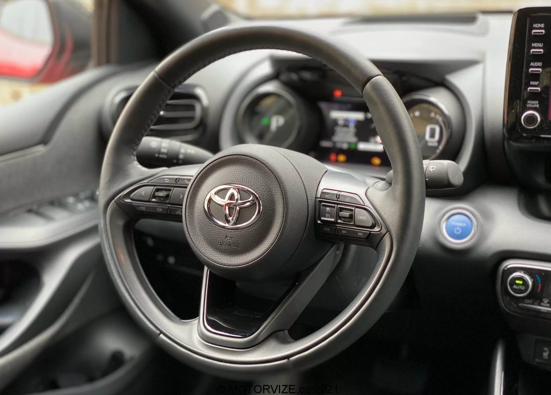 Vista interior de un Toyota Yaris 2020 enfocándose en el volante y el tablero desde la perspectiva del asiento del conductor. El volante presenta el logo de Toyota y controles visibles para el sistema de infoentretenimiento y sistemas de asistencia al conductor. Detrás de él, se pueden ver partes del panel de instrumentos digital y la consola central con controles de climatización y salidas de aire.