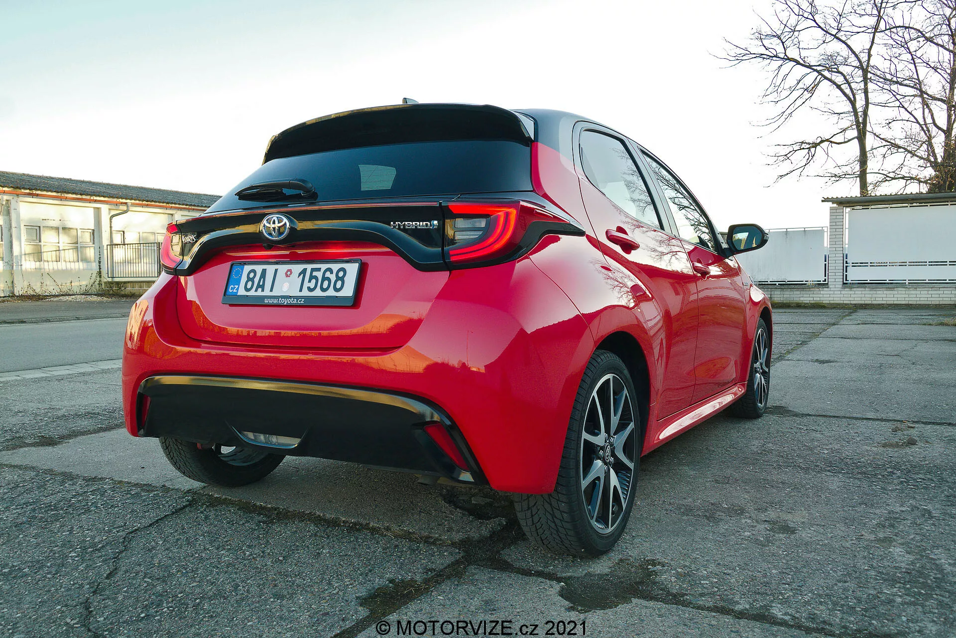 Vibrante coche de pasajeros Toyota Yaris 2020 rojo fotografiado desde un ángulo trasero de tres cuartos, destacando las luces traseras LED, el alerón trasero, el emblema del modelo híbrido, llantas de aleación de varios radios y características visibles de asistencia al conductor como los sensores de estacionamiento y la cámara trasera, capturado desde una perspectiva ligeramente inferior a la altura del coche.