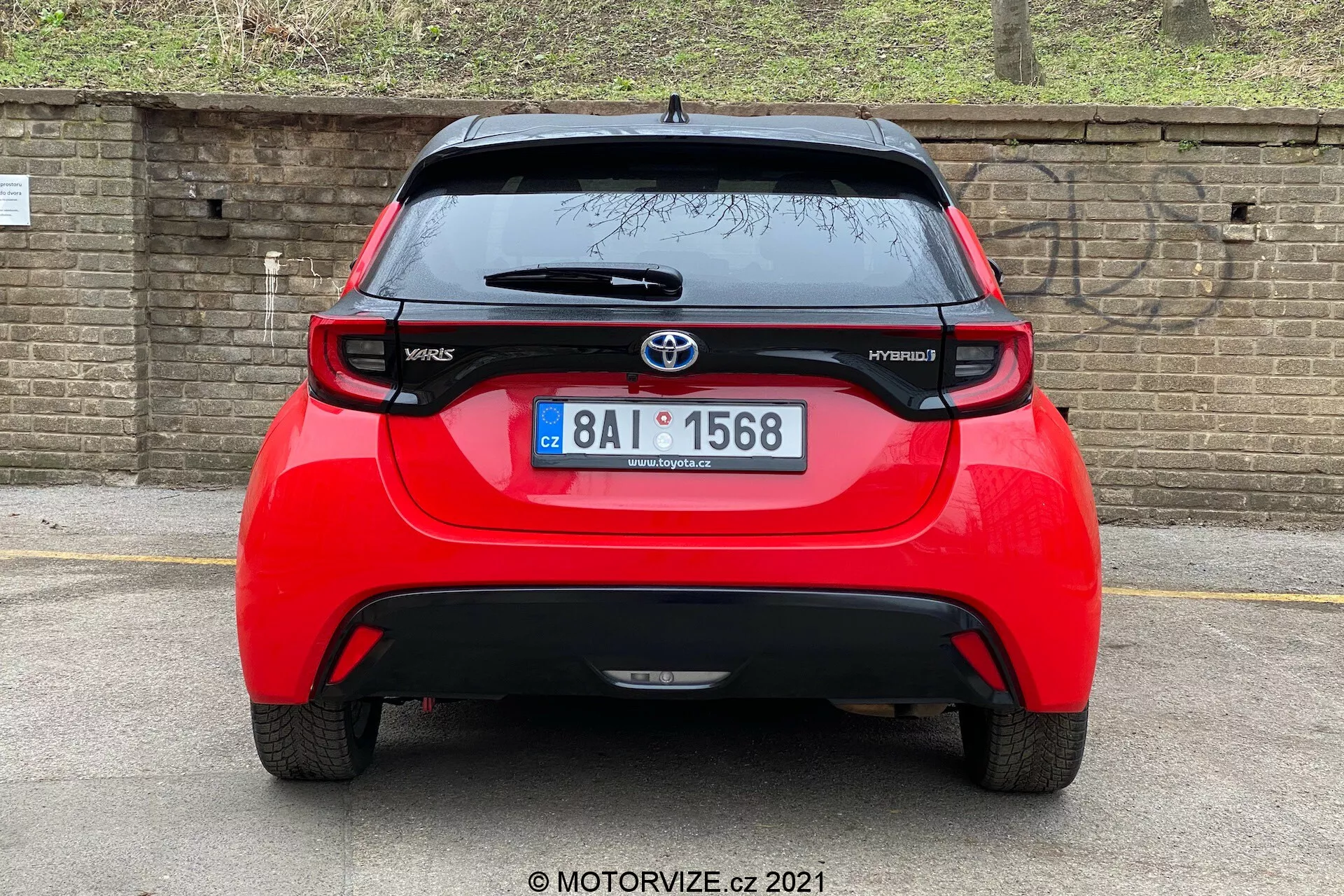 Vista trasera de un Toyota Yaris Hybrid rojo del 2020, centrada y tomada en un ángulo directo. La imagen revela todo el ancho del vehículo, mostrando el emblema de Toyota, los nombres de los modelos 'Yaris' y 'Hybrid', un alerón, una antena montada en el techo, una franja de luz de freno alta y luces traseras distintivas. También es visible la ventana trasera con un limpiaparabrisas y una parte baja del parachoques que probablemente albergue sensores o reflectores. El fondo incluye una pared de ladrillo o una cerca y algo de vegetación, lo que sugiere un coche aparcado más bajo o un ángulo de cámara más bajo.