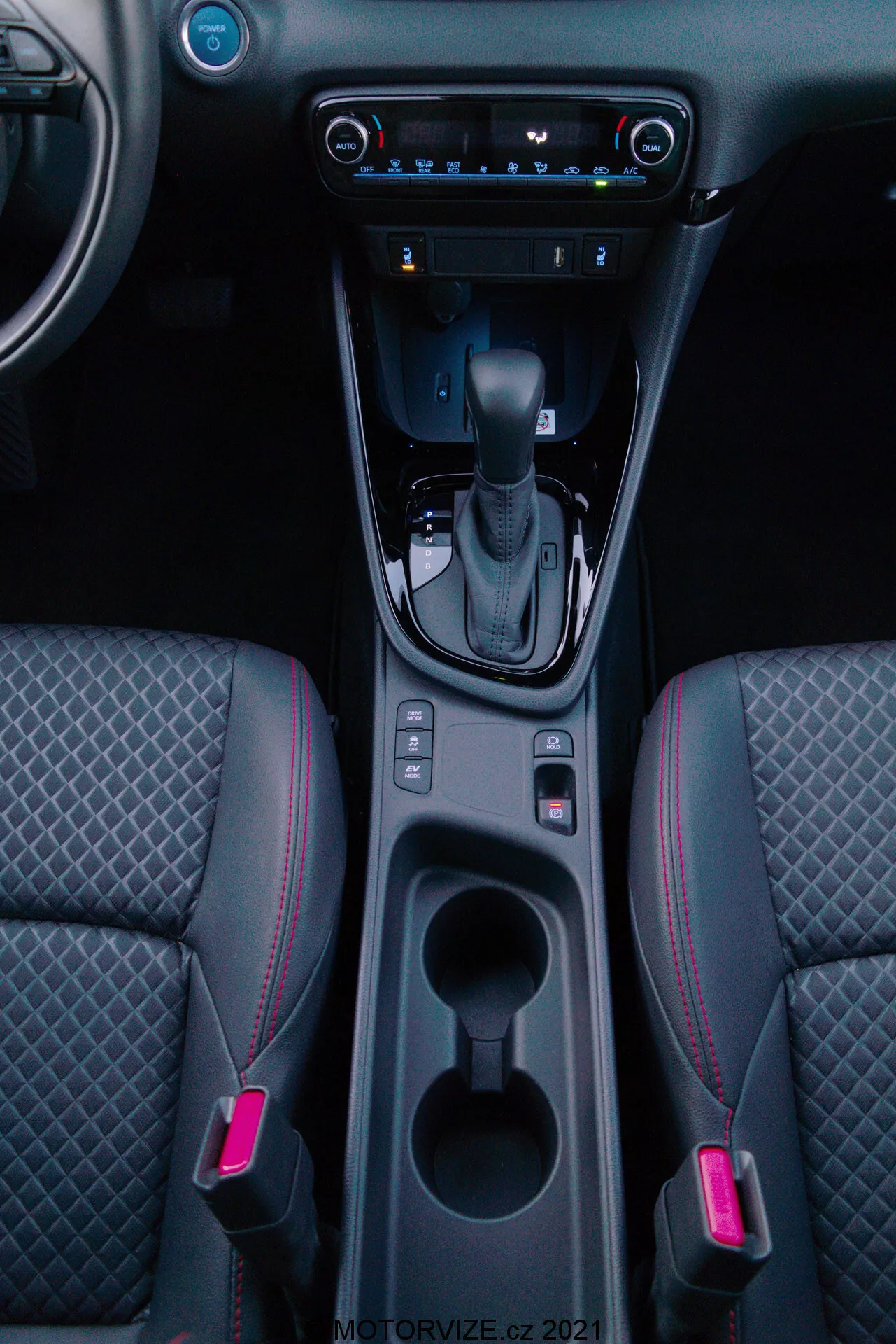 Imagen vertical del interior de un Toyota Yaris 2020 enfocada en la consola central, que presenta la palanca de cambios en posición 'P', botón de freno de estacionamiento electrónico, dos portavasos iluminados, espacio de almacenamiento, control de clima digital, sistema multimedia de pantalla táctil, puerto USB, salidas de aire y asientos delanteros bicolor con costuras en patrón de diamante y costuras de acento rojo.