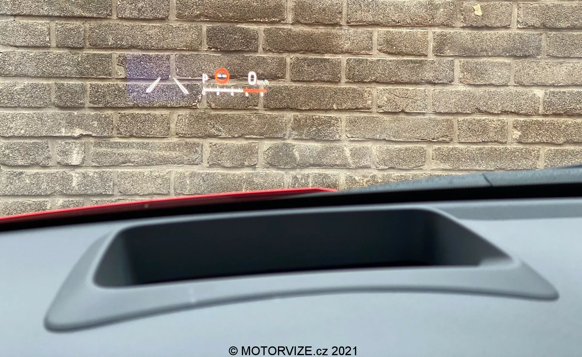 Vista de cerca desde el interior de un Toyota Yaris 2020 mostrando una perspectiva parcial desde el asiento del conductor hacia la parte trasera. El enfoque está en el estante trasero con una luz de freno montada en alto y un alerón trasero integrado. La imagen también muestra parte del material interior mientras que una pared de ladrillo afuera sugiere que el coche está estacionado cerca de un edificio.