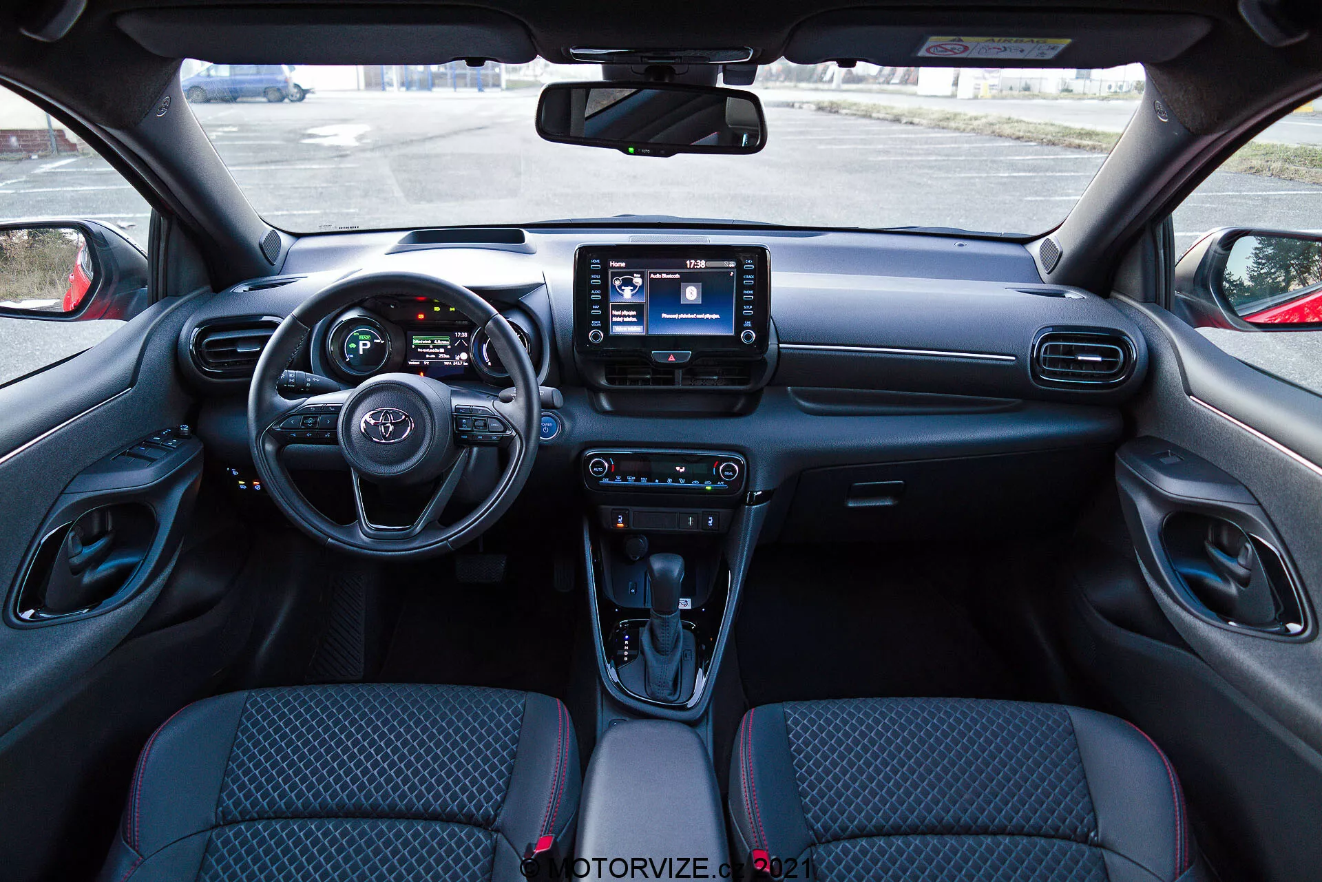 Vista del interior de un Toyota Yaris 2020 desde la perspectiva del asiento del conductor, con un tablero moderno que incluye un volante con controles montados, un cuadro de instrumentos parcialmente visible con pantallas digitales y un sistema de infoentretenimiento central con interfaz de pantalla táctil. El tablero cuenta con ventilaciones de aire y botones de control climático, mientras que la consola central tiene controles para modos de manejo, una palanca de cambios automática con funda tipo cuero, portavasos y un compartimento de almacenamiento. Los asientos de tapicería de tela tienen un diseño estampado y los paneles interiores de las puertas incluyen controles para ventanas y espejos eléctricos.