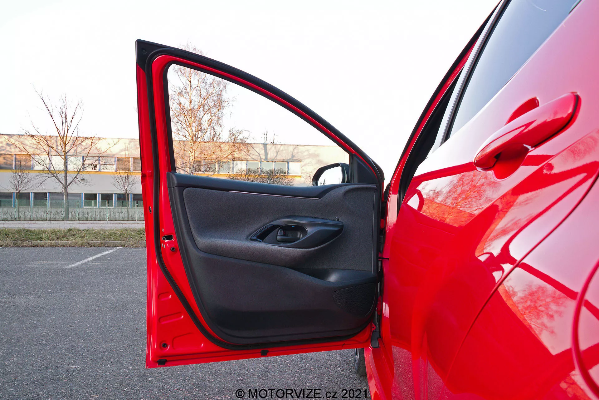 Vista en primer plano del lado del conductor de un Toyota Yaris 2020 rojo con la puerta delantera abierta, capturada desde un ángulo ligeramente bajo. La cámara está posicionada afuera, cerca de la puerta, apuntando hacia el interior, mostrando la pintura exterior roja brillante que refleja la luz y el adorno interior negro con un diseño minimalista. La puerta tiene un botón de control de ventana eléctrica dentro del hueco de la manija y un bolsillo de almacenamiento debajo.